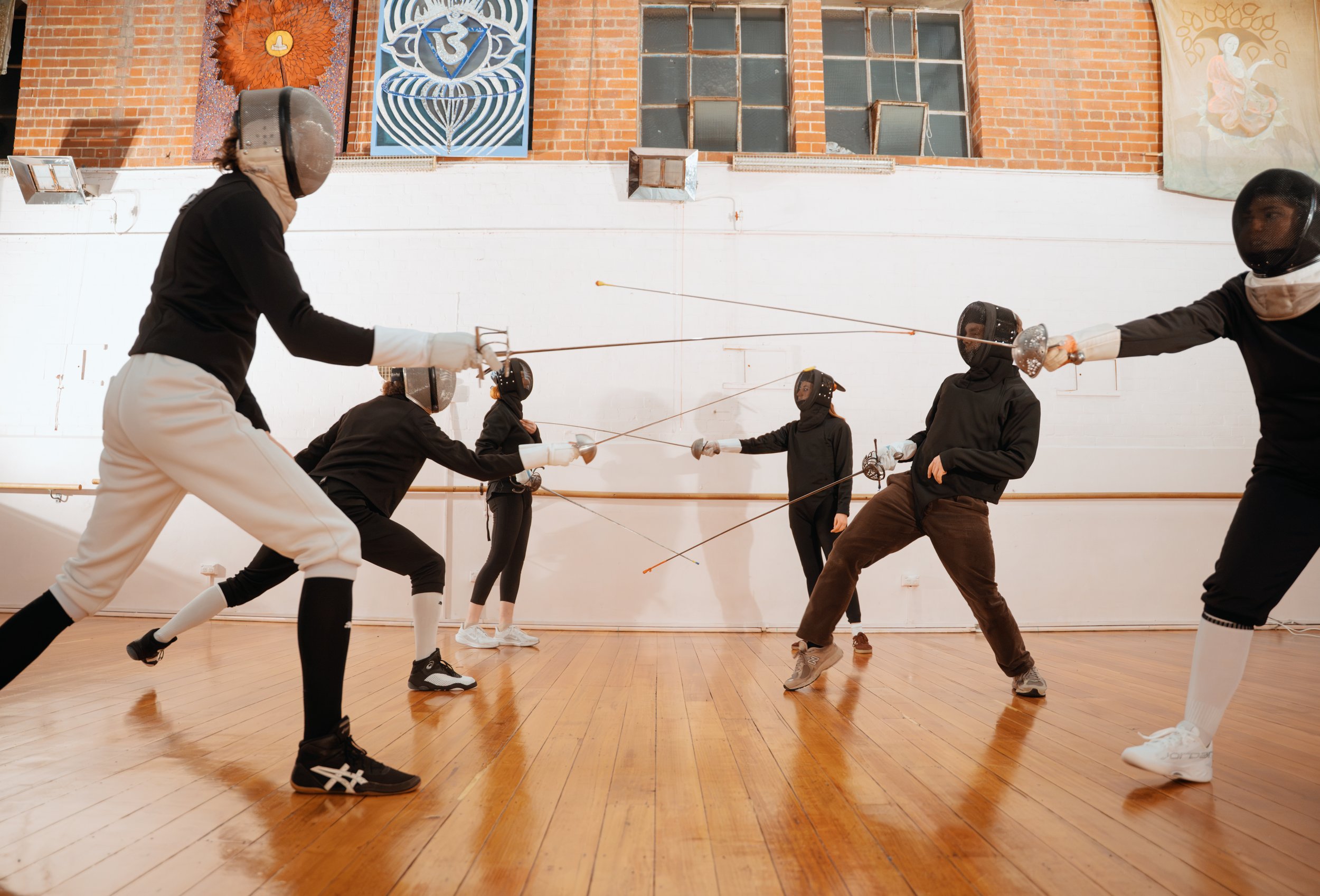 Group of people practicing fencing in a gymnasium with wooden floors and large windows, some wearing fencing masks and holding swords.