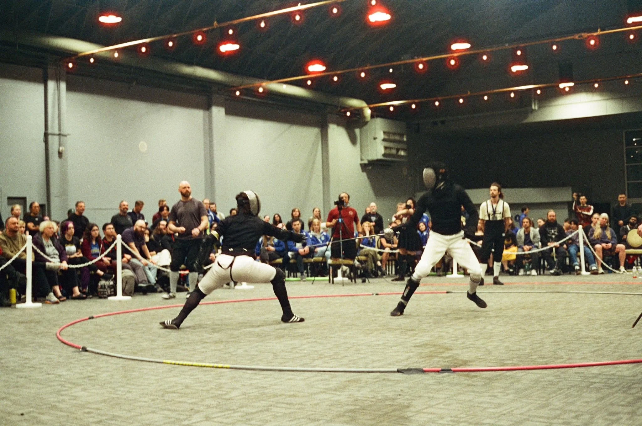 Two fencers are fencing at a high profile tournament, with a large spectator audience watching, seated behind a rope barrier.