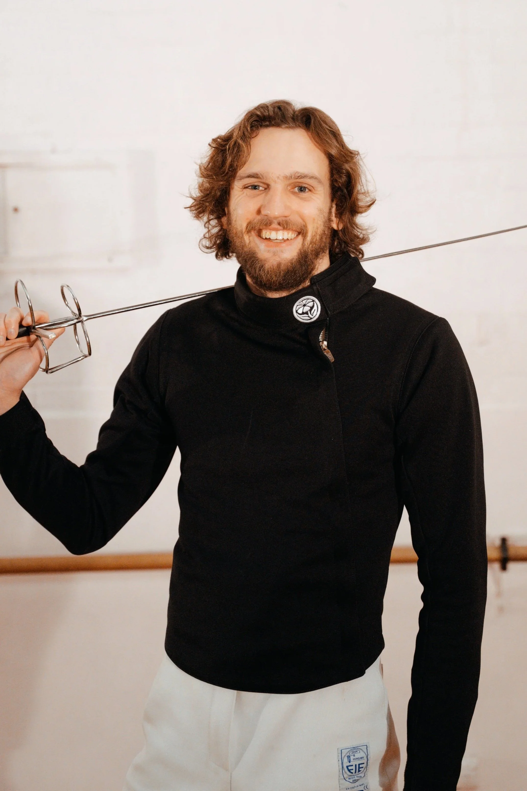 Head Instructor Woody Craig of Melbourne Historical Fencing is smiling with his Rapier against a white wall.