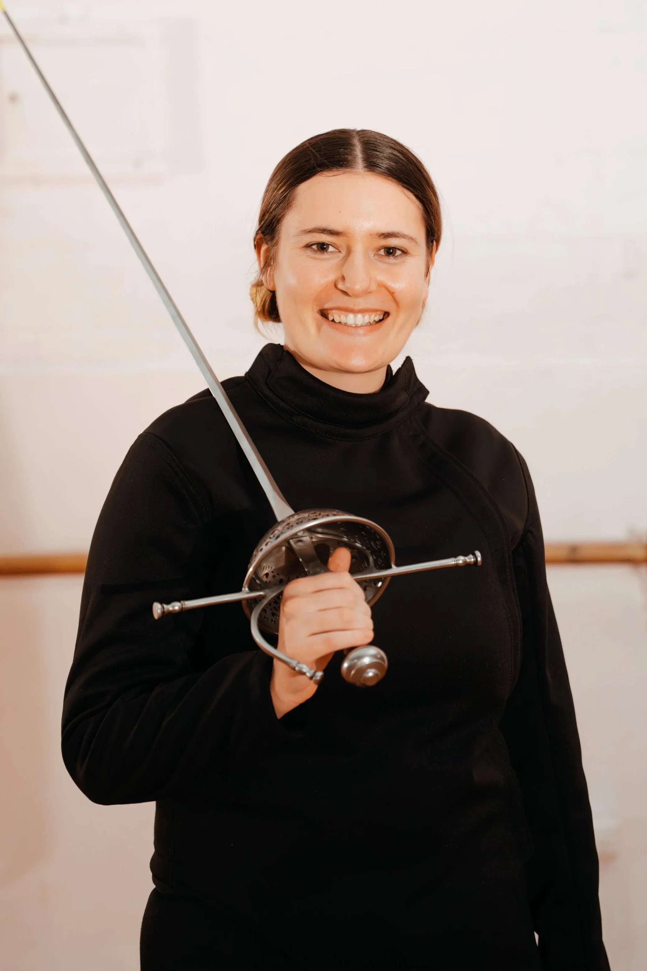 Operations Manager Heather Barry of Melbourne Historical Fencing is smiling with her Rapier against a white wall.