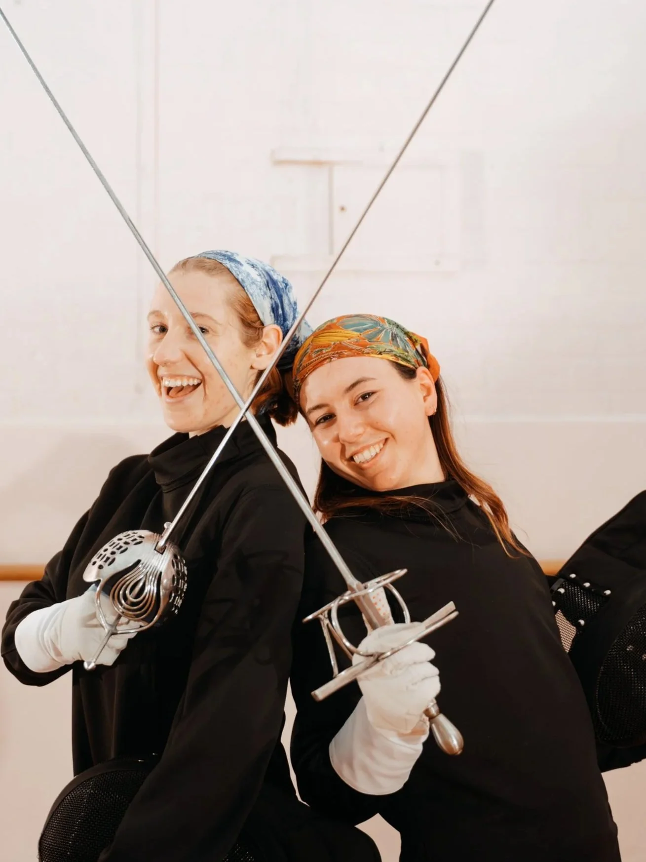 Two women wearing brightly coloured headscarves are posing while laughing, holding their swords crossed