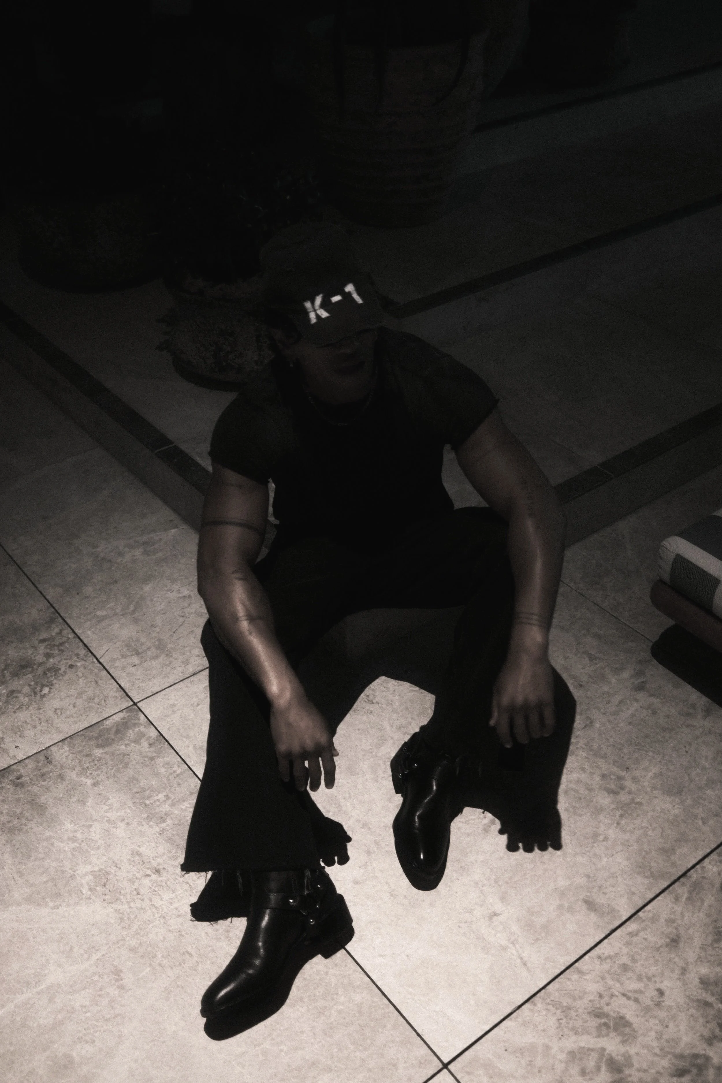 Person in dark clothing and black boots sitting on tiled floor in a dimly lit room with patterned ceiling and black balloons.