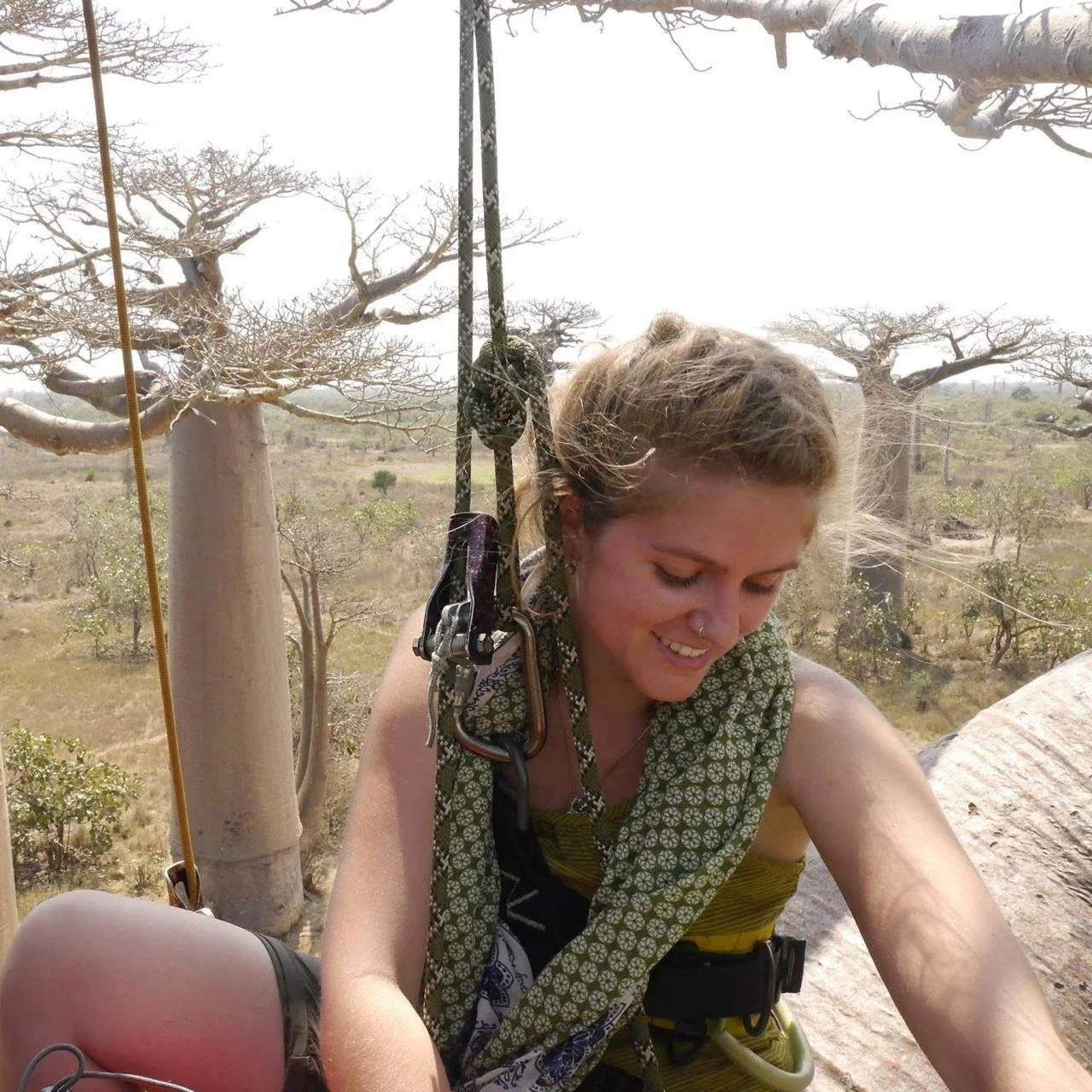 A woman with blonde hair and a nose ring smiling in a harness during a climbing activity in a dry, desert-like landscape with leafless trees.