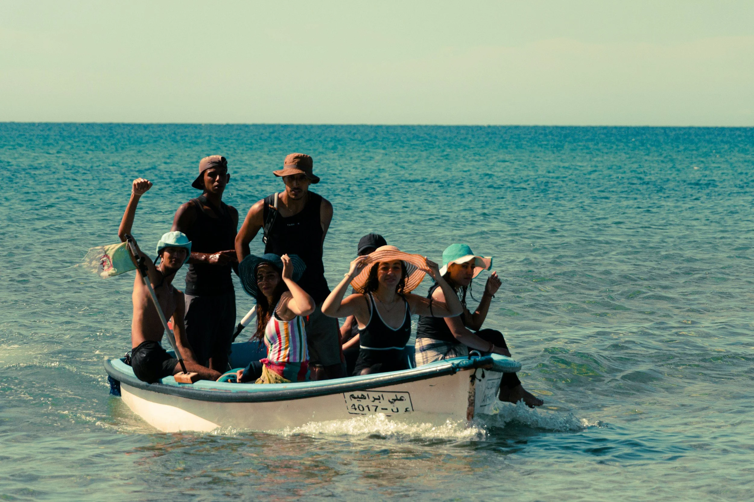 A group of people on a boat in the water at the beach, some wearing hats and swimsuits, with the ocean and clear sky in the background.