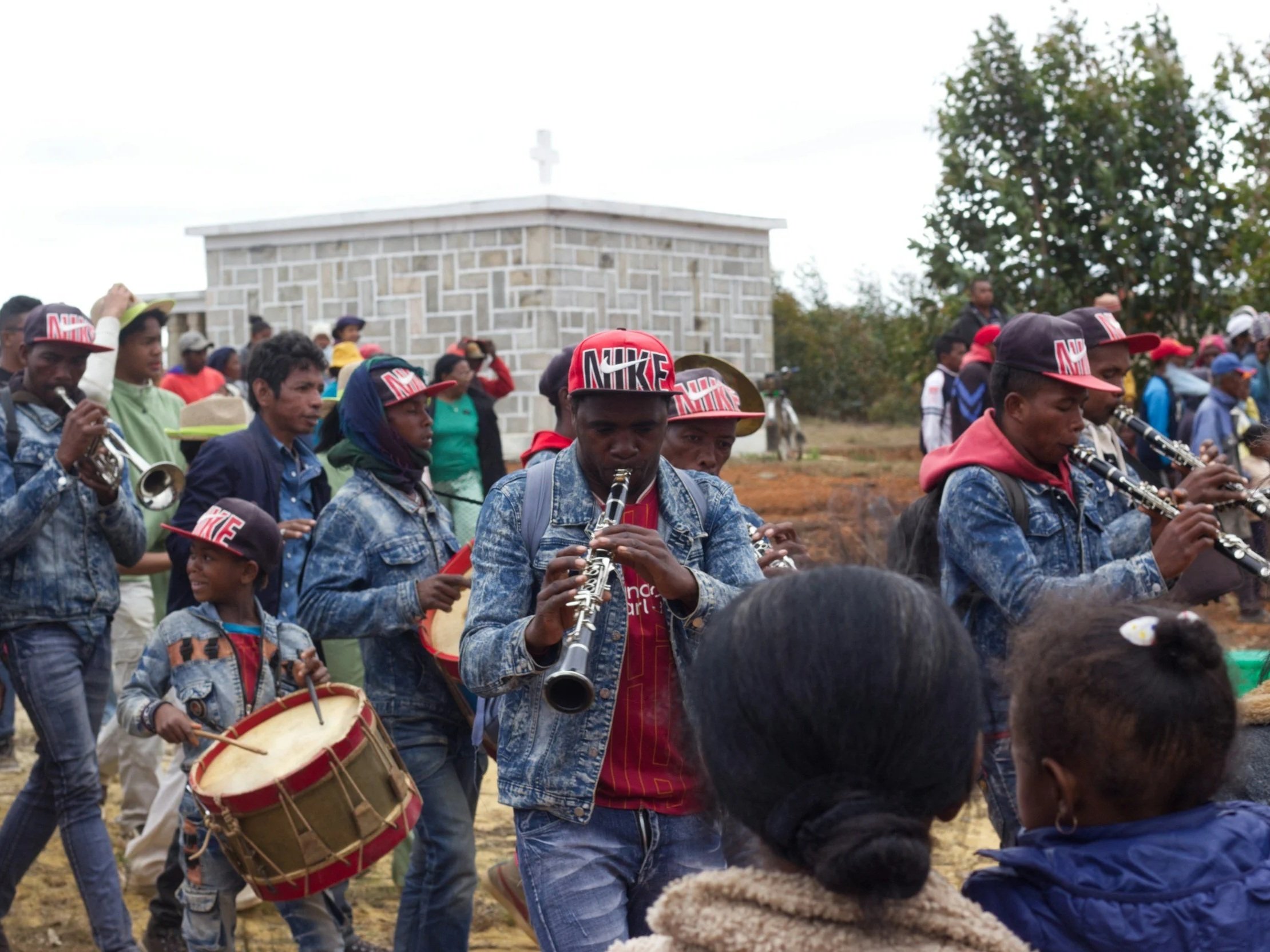 A group of people playing musical instruments outdoors, some wearing Nike caps, near a stone building with a white cross on top and trees in the background.