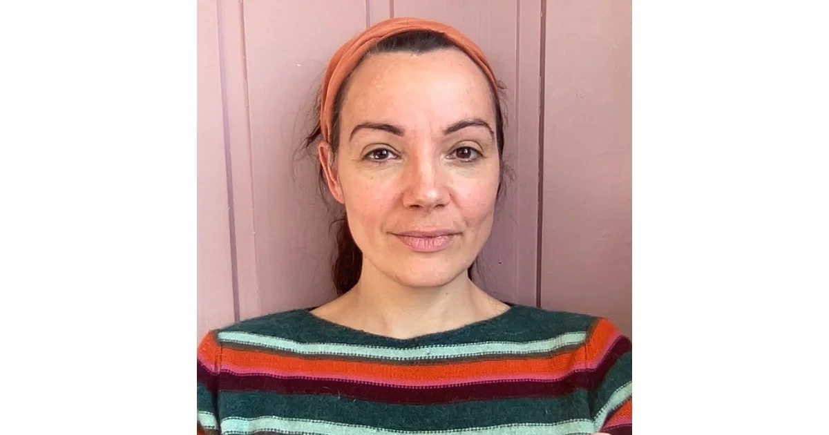A woman with a peach headband, wearing a multicolored striped sweater, standing in front of a pink wall.