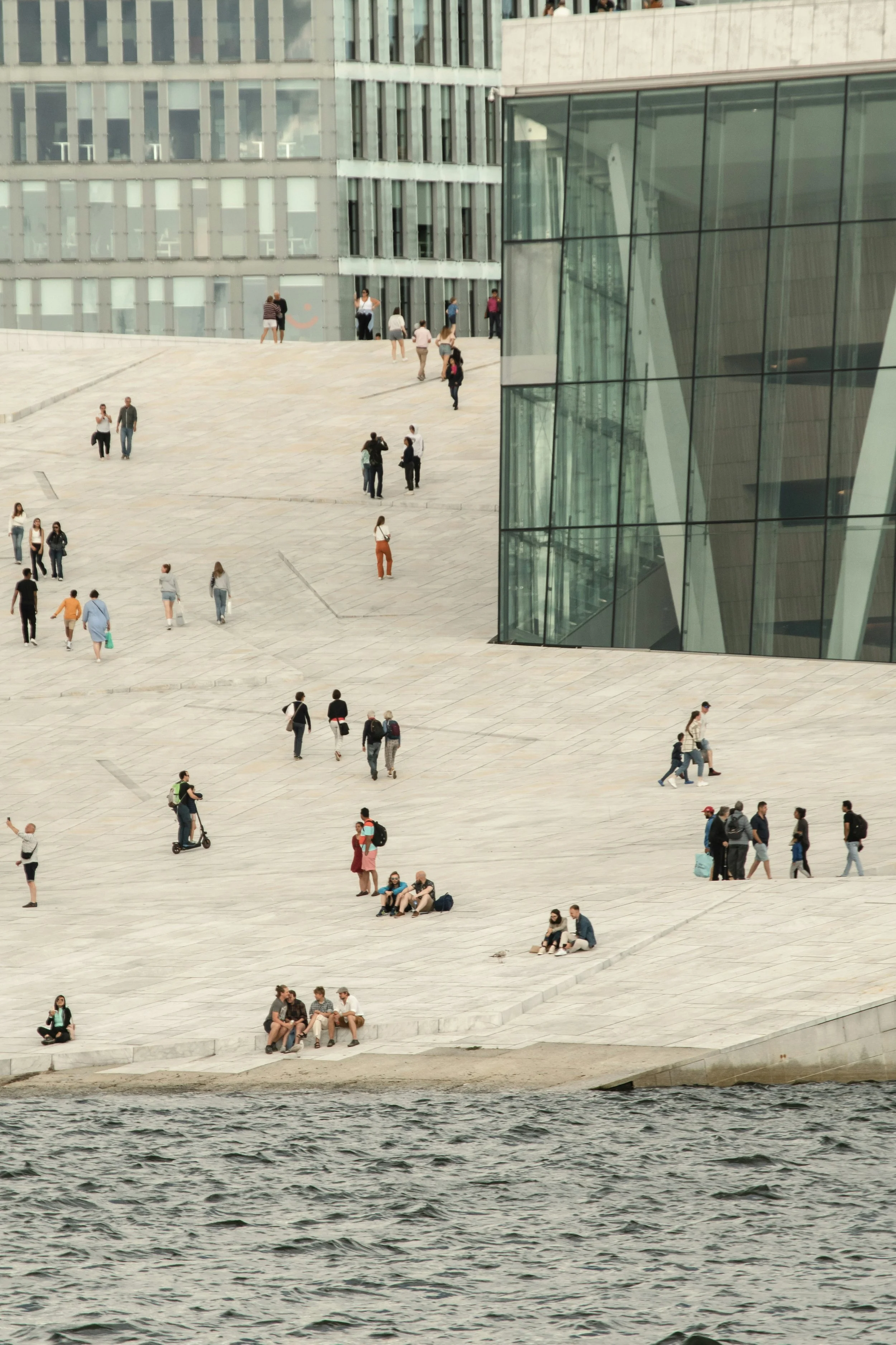 People walking, sitting, and socializing on a large marble staircase near a waterfront in an urban area, with modern glass buildings.
