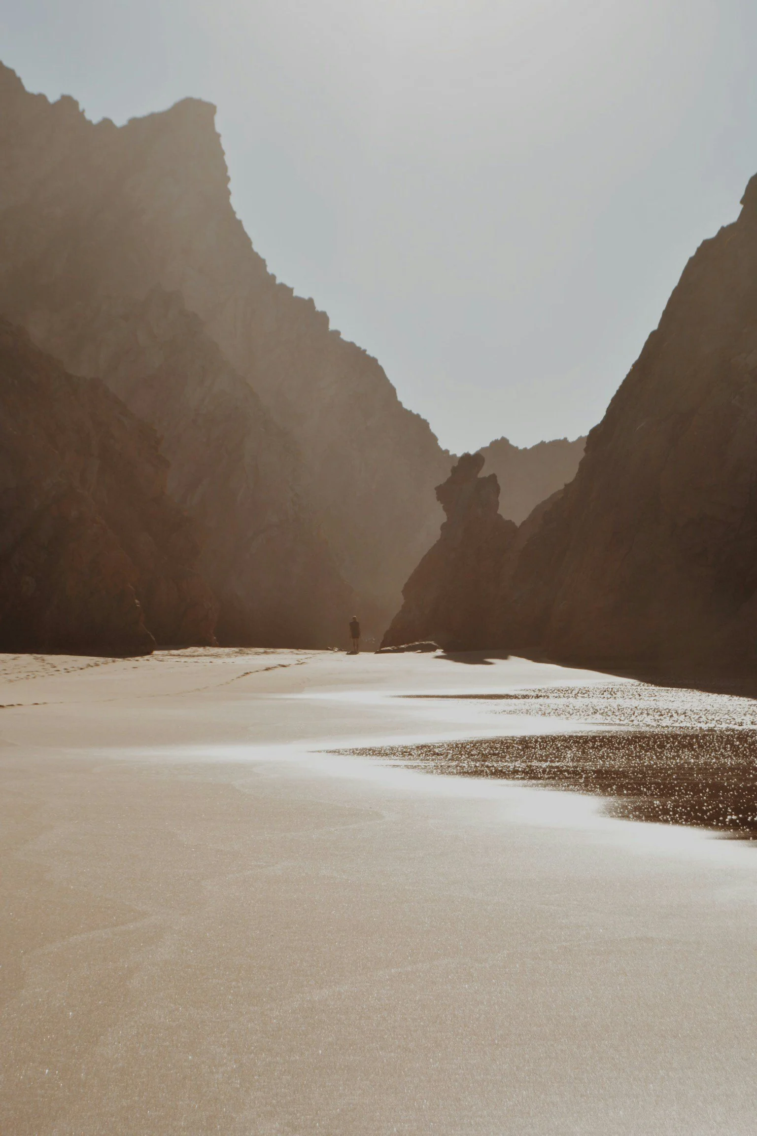 A person standing alone on a beach surrounded by tall rocky cliffs, with the sandy shore extending in the foreground and a hazy sky overhead.