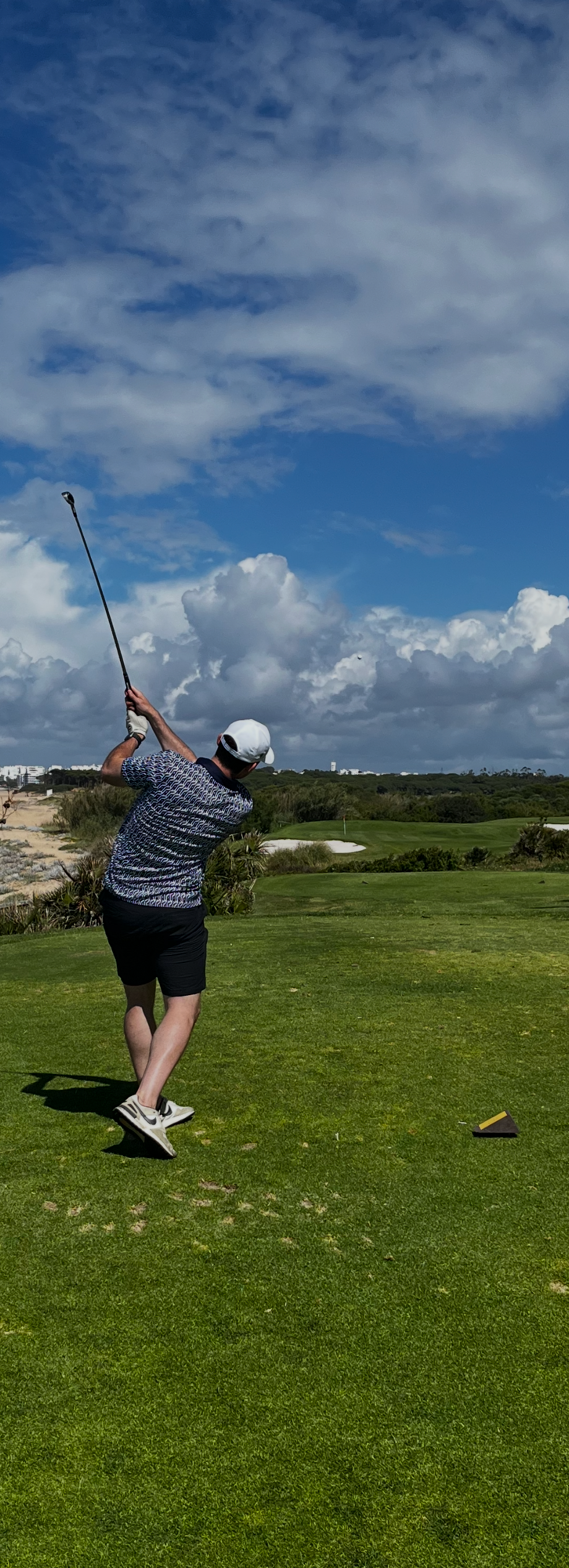 A man playing golf on a course, swinging a golf club under a partly cloudy sky.