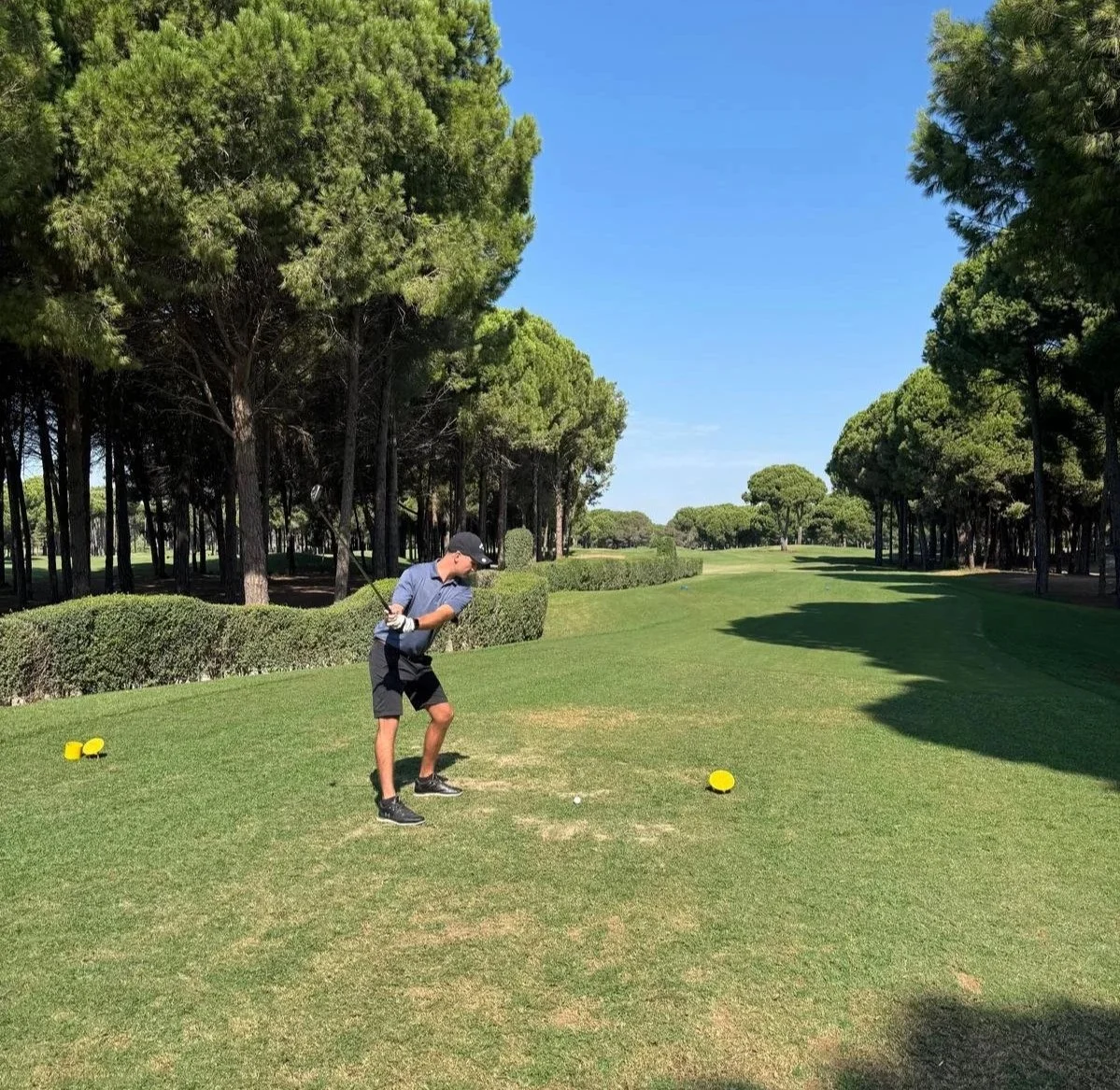 A young golfer in a blue shirt, black shorts, and a cap preparing to hit a golf ball on a lush green golf course with trees and a clear blue sky in the background.
