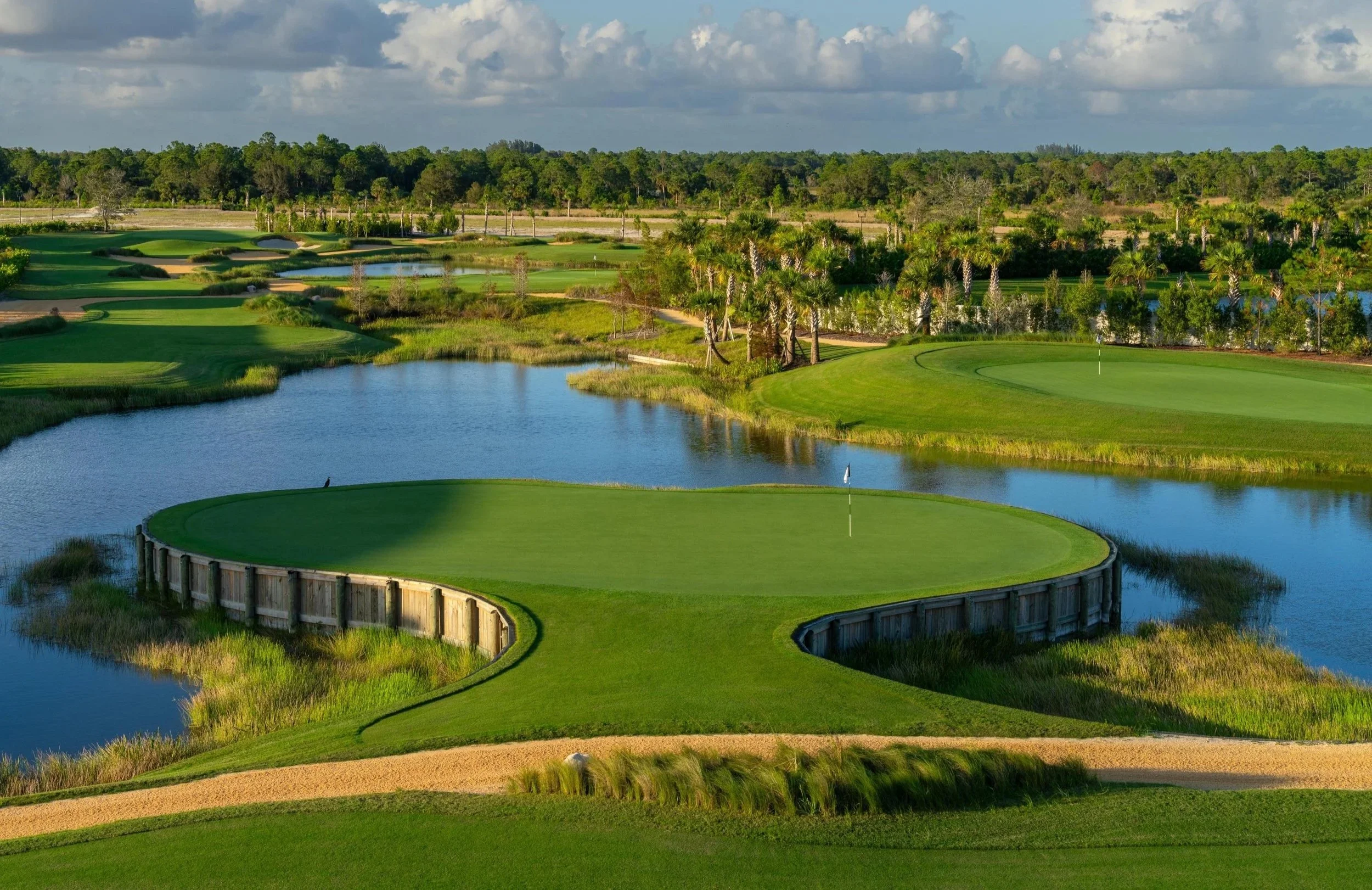 A view of a golf course with a green surrounded by a water hazard and sand trap, bordered by trees and a wooden fence in a sunny landscape with clouds in the sky.