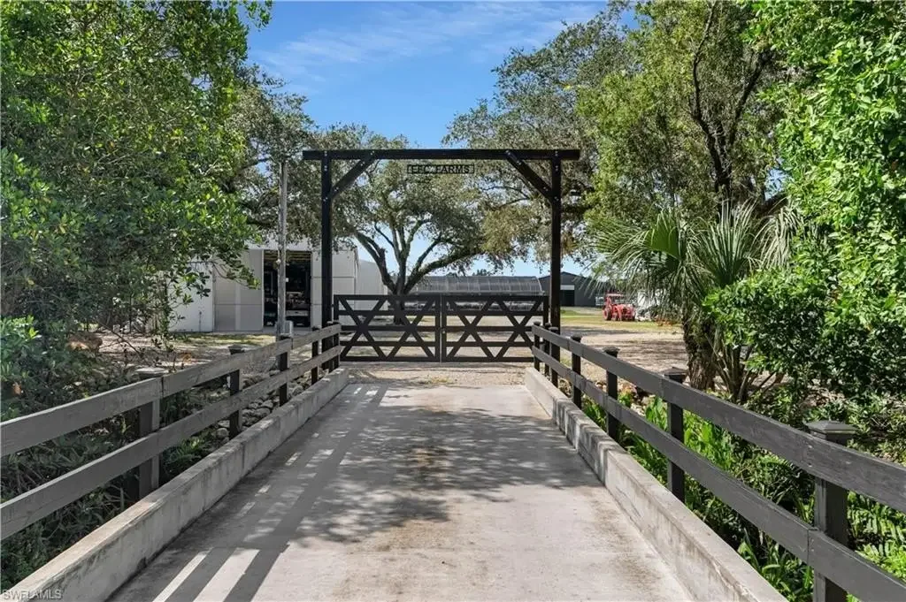 A wooden bridge with black railings leading to a farm gate, surrounded by greenery and trees, with a partly cloudy blue sky overhead.