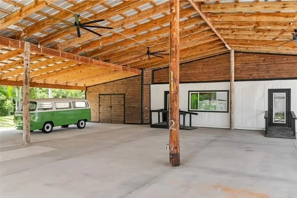 Open wooden structure with a cement floor, ceiling fans, and a small green and white vintage van inside. The structure has a rustic wooden roof and support beams, with a building wall featuring windows and doors visible.