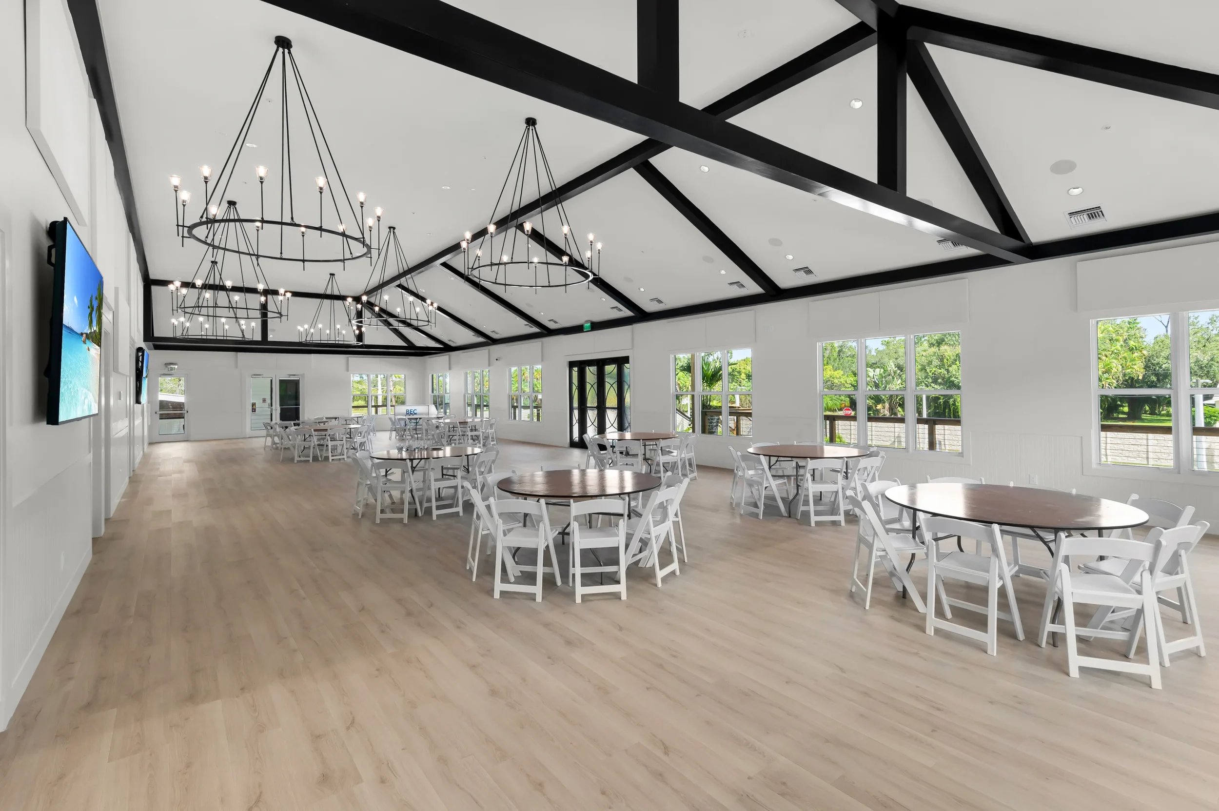 Empty banquet hall with white walls, large windows, wooden flooring, round tables, white chairs, and black chandeliers hanging from a white ceiling with black beams.