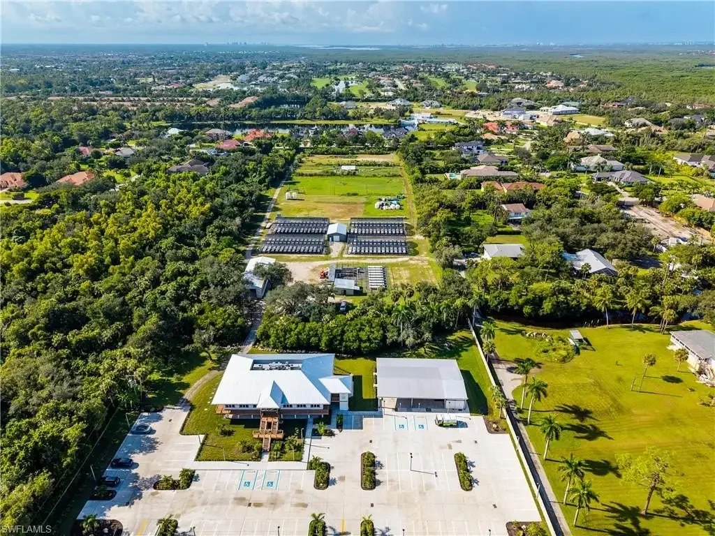 Aerial view of a community featuring a building with a white roof, a parking lot with designated handicapped spots, surrounded by lush trees and grassy areas. In the background, there are residential houses and green fields extending toward the horizon.