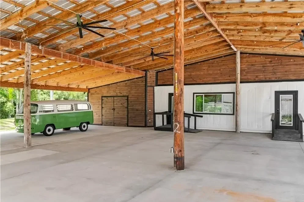 Inside a spacious, open garage or barn with a corrugated metal roof, wooden support beams, and ceiling fans. There is a green vintage Volkswagen bus parked on concrete flooring, and a small wooden building with a window, door, and ramp for accessibility.