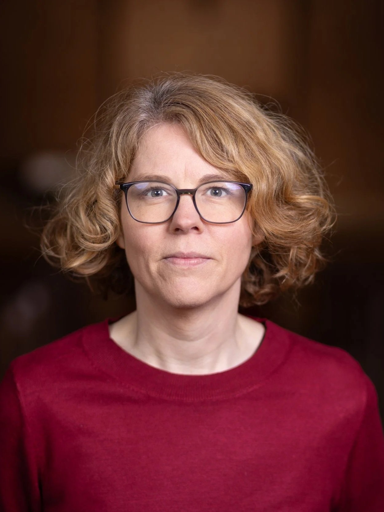 Porträt eines Frau mit Locken, Brille und rotem Pullover vor dunklem Hintergrund.