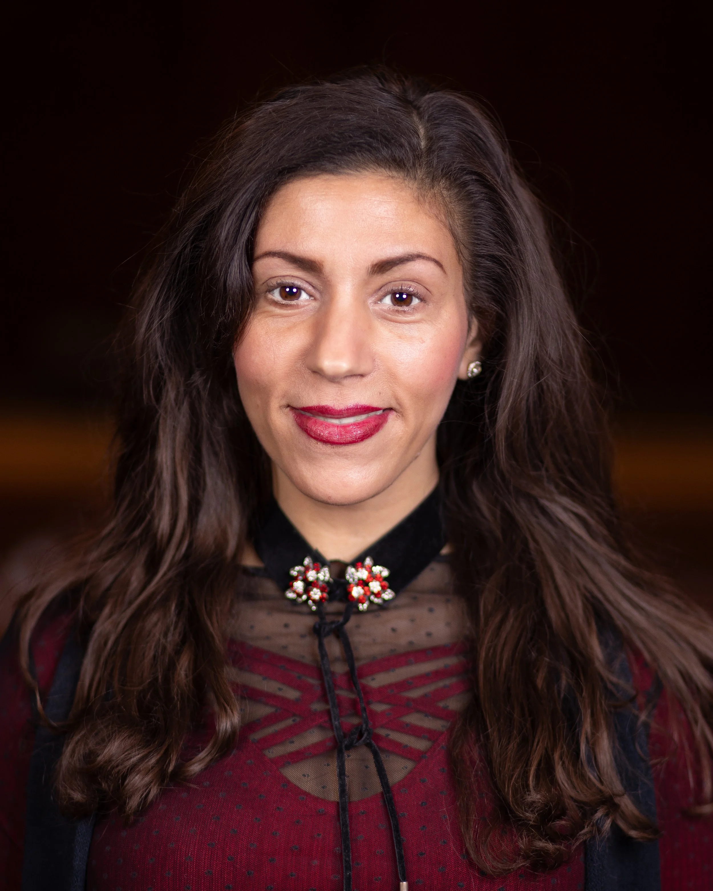 Nahaufnahme einer Frau mit dunklen, lockigen Haaren, roten Lippen und einem schwarzen, frauenfreundlichen Kleid mit roten und schwarzen Streifen, sowie einer dekorativen Halskette.