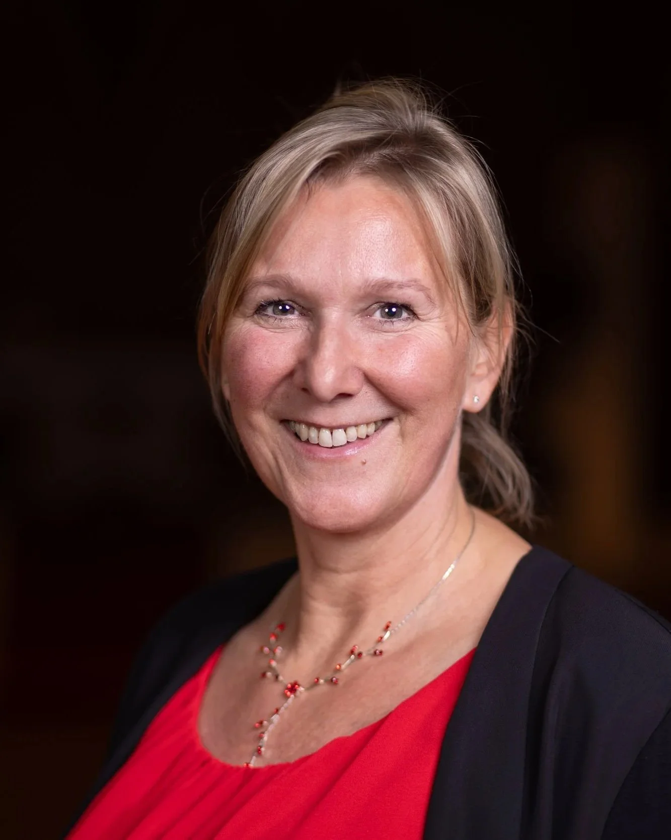 Portrait einer lächelnden Frau mit blonden Haaren, schwarz-rot Kleidung, kleinen Ohrringen und einer roten Halskette vor dunklem Hintergrund.