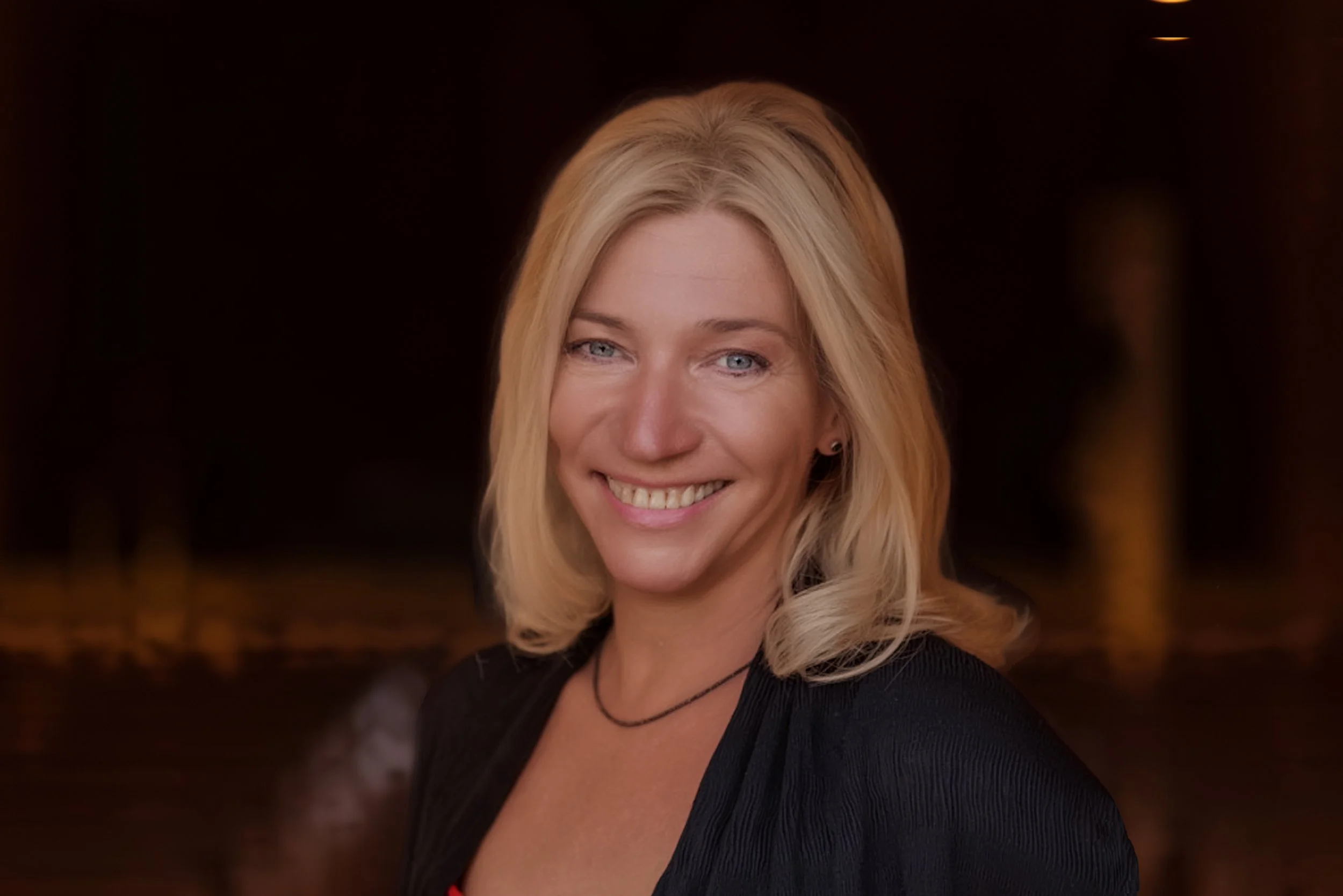 Porträt einer lächelnden Frau mit blonden, lockigen Haaren gegen einen dunklen Hintergrund.