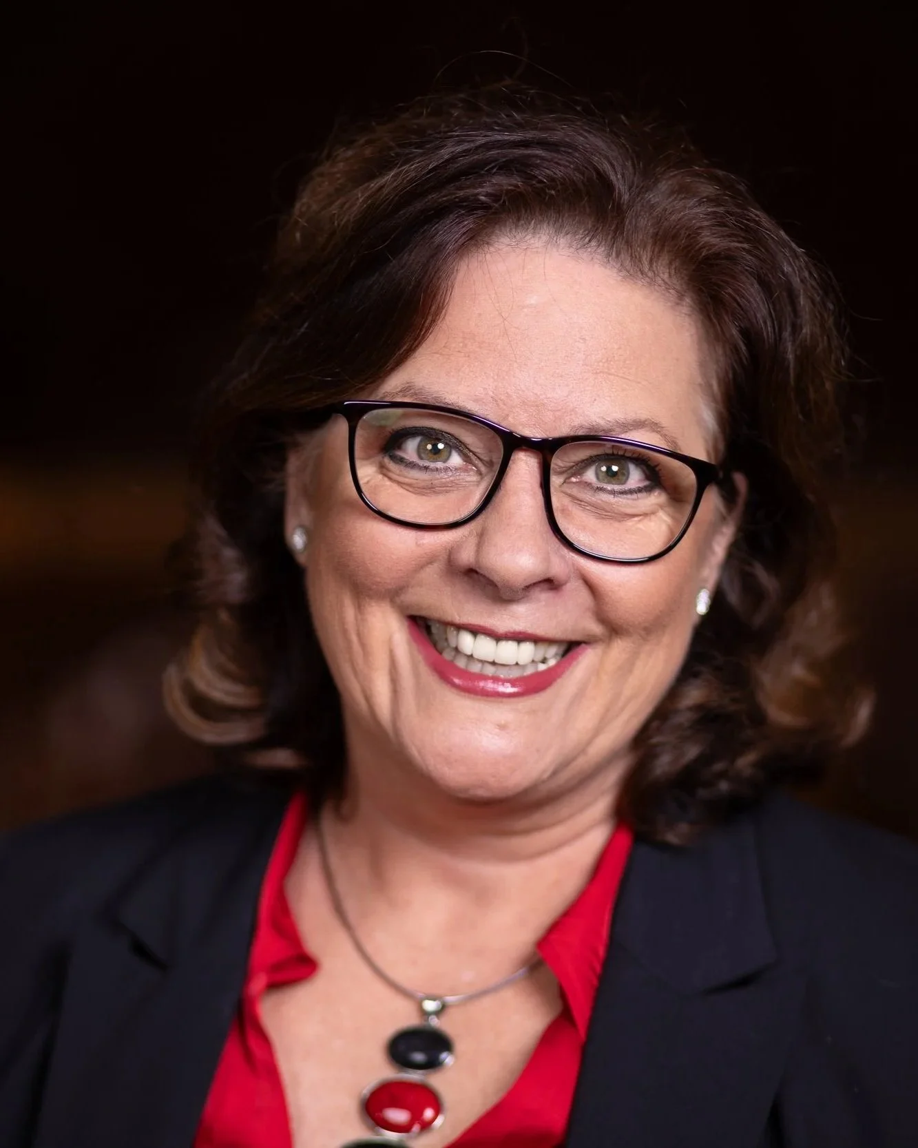 Eine lächelnde Frau mit Brille, braunen Haaren und rotem Lippenstift, trägt eine schwarze Jacke, eine rote Bluse und eine silberne Halskette.