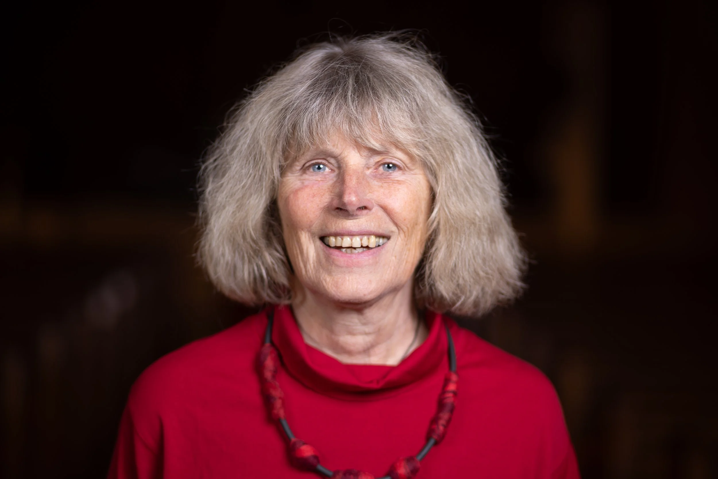 Porträt einer Frau mit grauem, lockigem Haar, blauen Augen und einem freundlichen Lächeln, trägt eine rote Bluse und eine rote Lederkette vor dunklem Hintergrund.