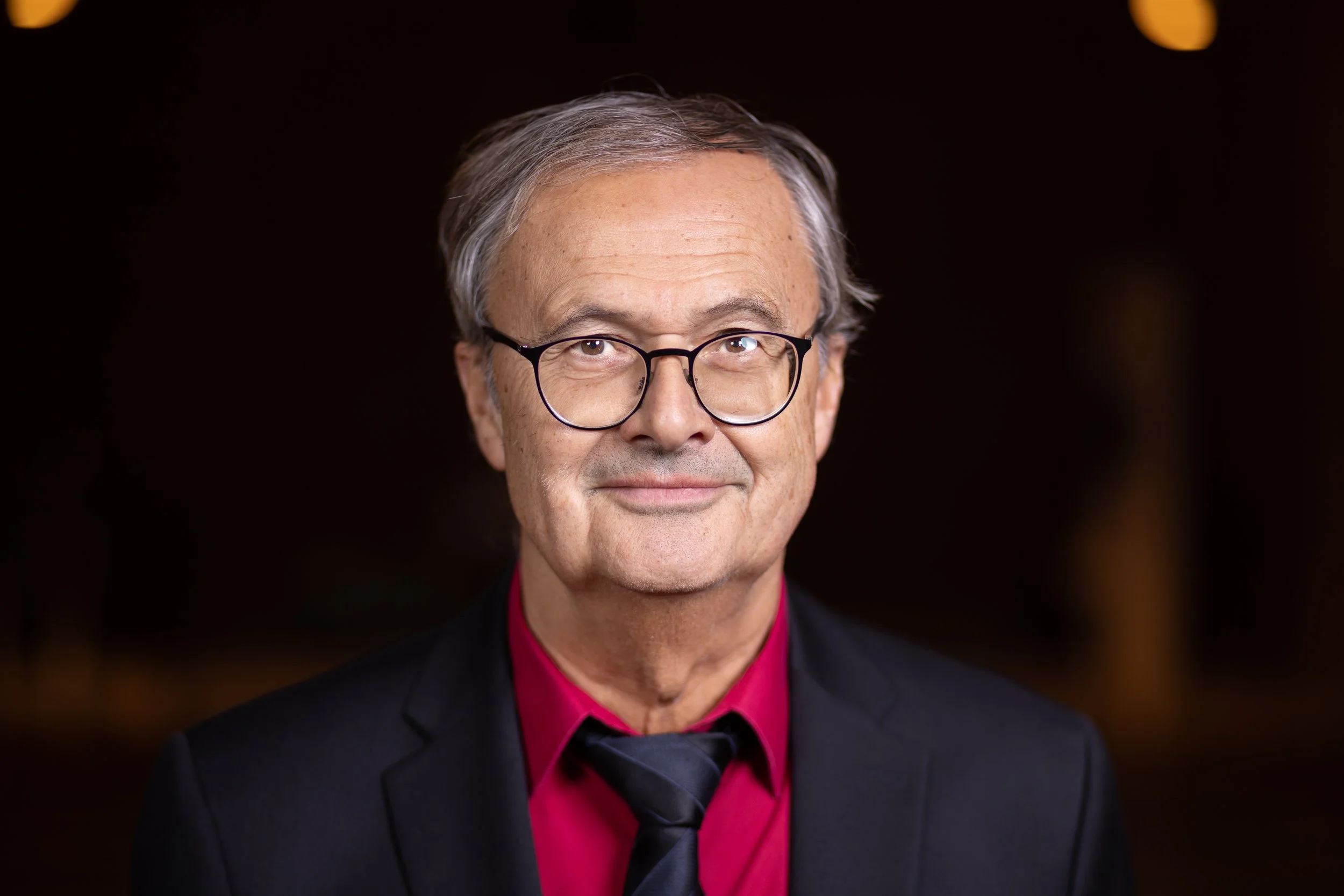 Porträt eines Mannes mit grauen Haaren, Brille, schwarzem Anzug, rotem Hemd und schwarzer Krawatte vor dunklem Hintergrund.