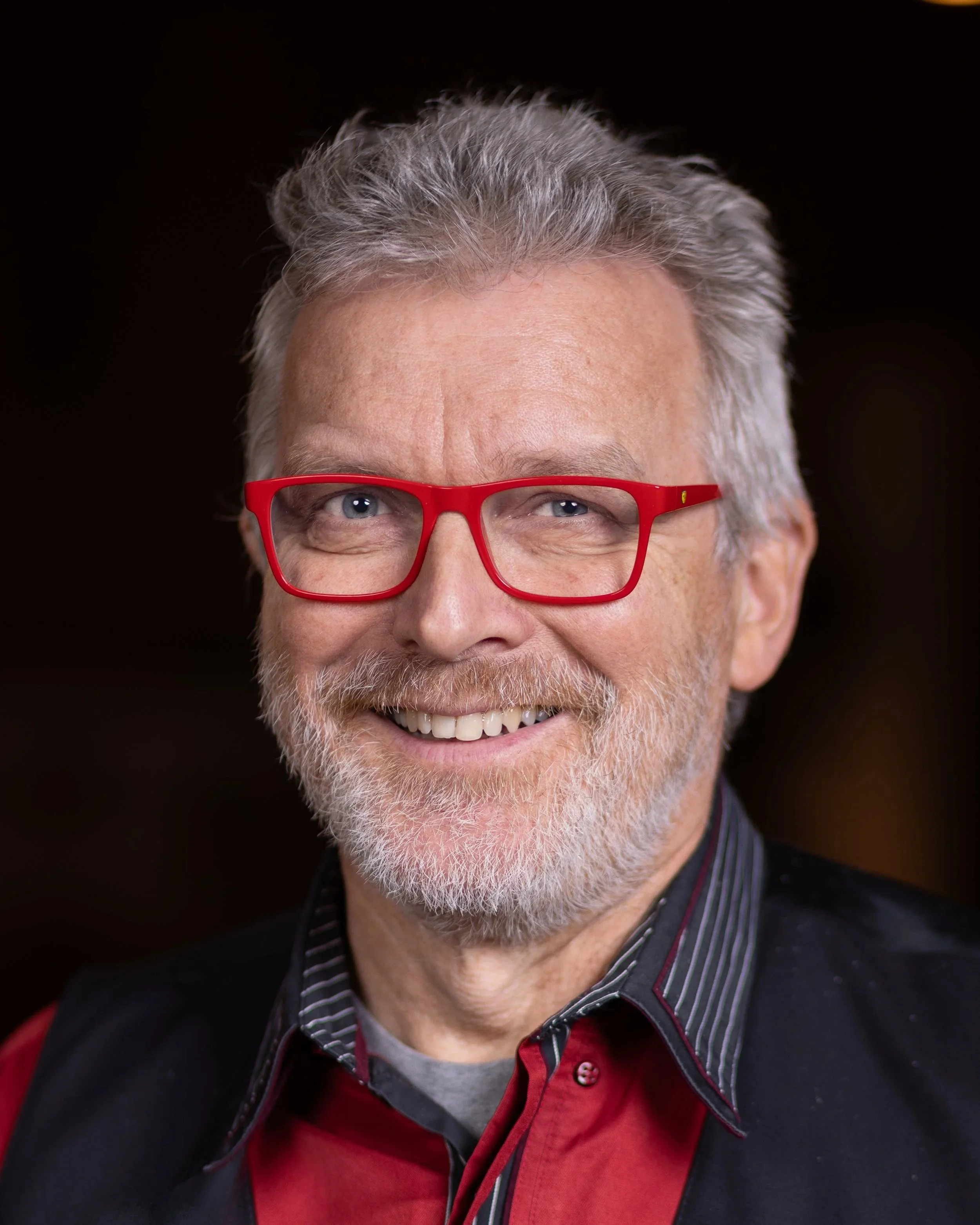 Nahaufnahme eines Mannes mit grau-weißem Haar und Bart, trägt rote Brille und sieht lächelnd in die Kamera.