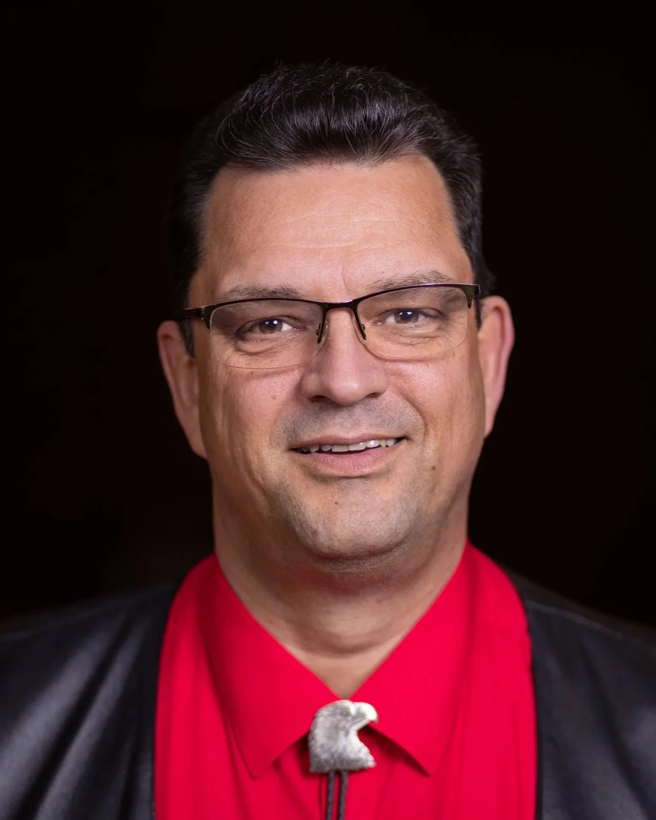 Porträt eines Mannes mit dunklen Haaren, Brille und rotem Hemd, gegen schwarzen Hintergrund.