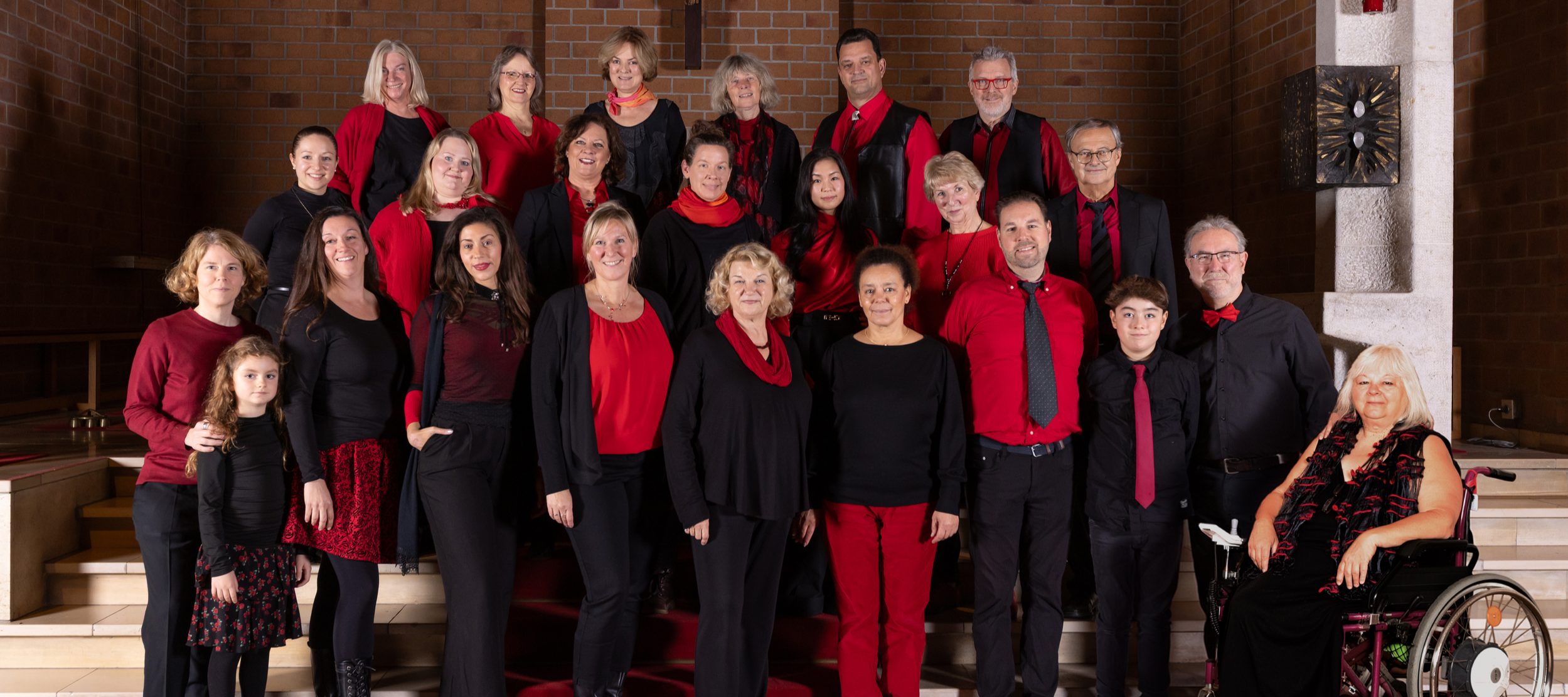 Gruppenfoto von mehreren Menschen, die in einem Kirchenraum stehen, alle tragen schwarze oder rote Kleidung, einige tragen rote Accessoires, und sie posieren für die Kamera auf Stufen vor einer Kirche.
