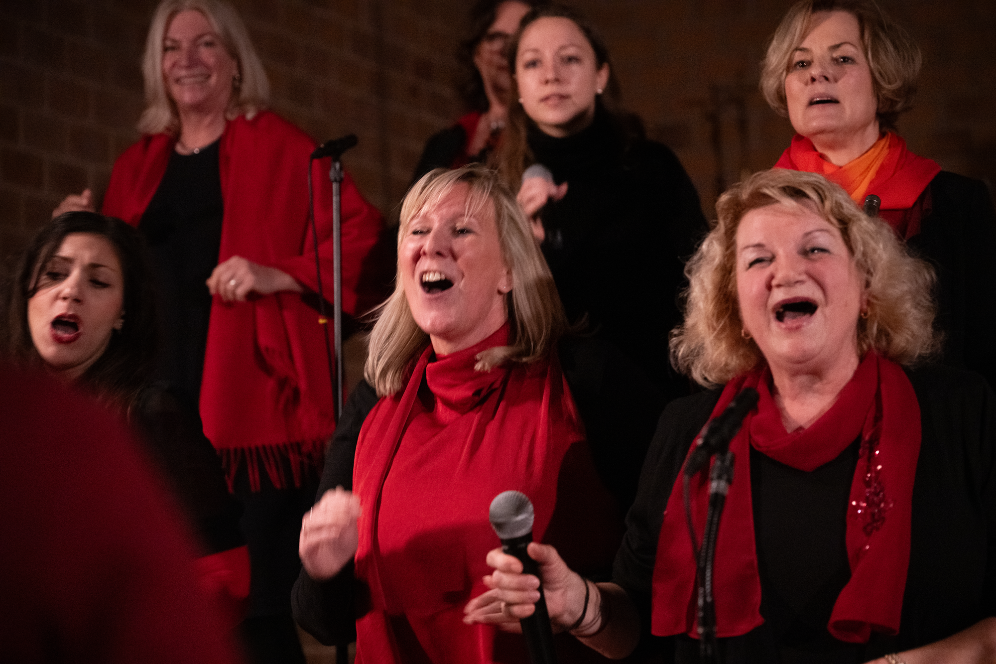 Eine Gruppe von Frauen singt und hält Mikrofone in einer Umgebung mit roten und schwarzen Kleidern, einige tragen rote Tücher.