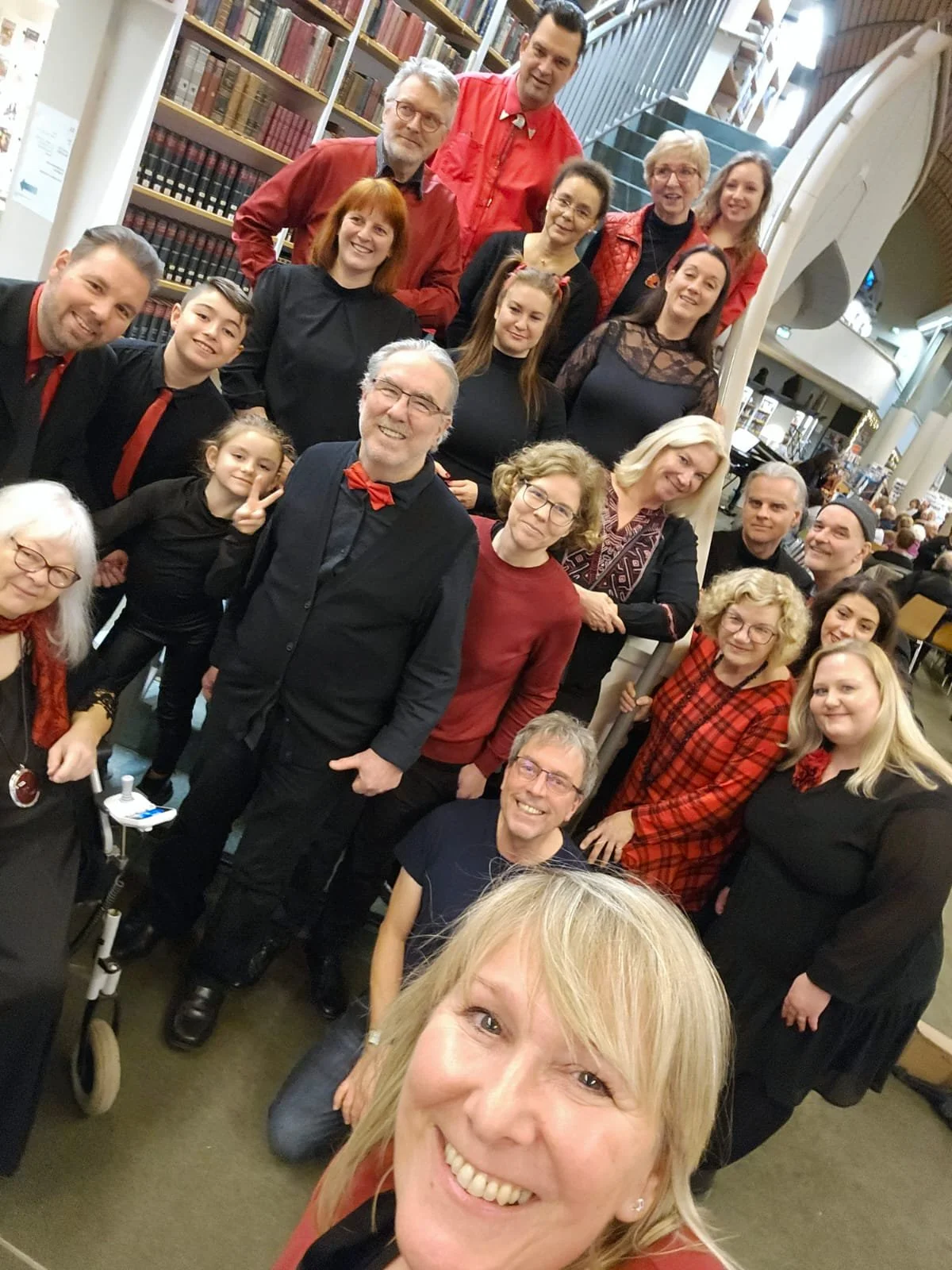 Gruppenfoto von mehreren Menschen, die in einer Bibliothek posieren, viele tragen rote und schwarze Kleidung, einige sind lächelnd und machen Handzeichen.