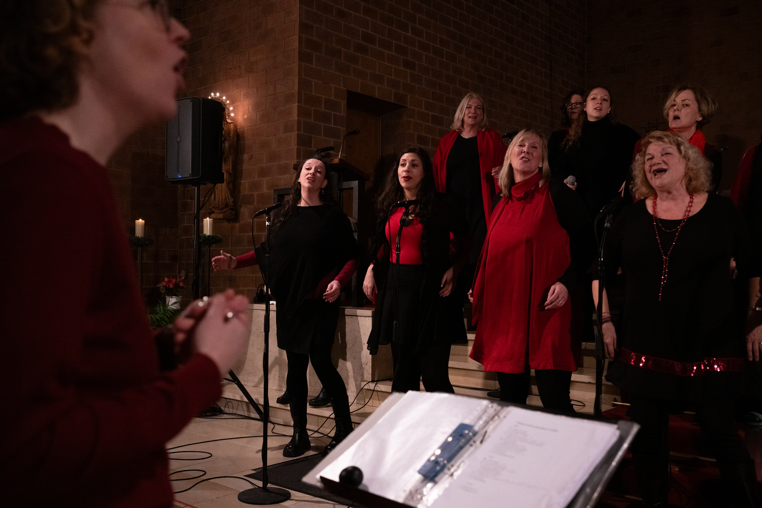 Mehrere Frauen singen in einem Raum mit Backsteinwänden, Kerzen und Weihnachtsdekoration.