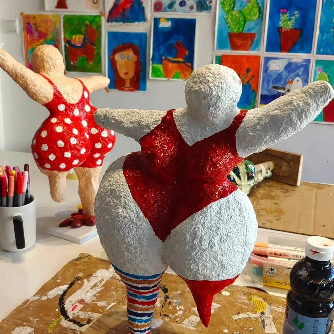Sculptures en papier mâché de deux personnages féminins, l'un avec un maillot de bain rouge à pois blancs, et l'autre avec un vêtement rouge et des chaussettes rayées, sur une table avec des outils artistiques et des œuvres d'art colorées en arrière-plan.
