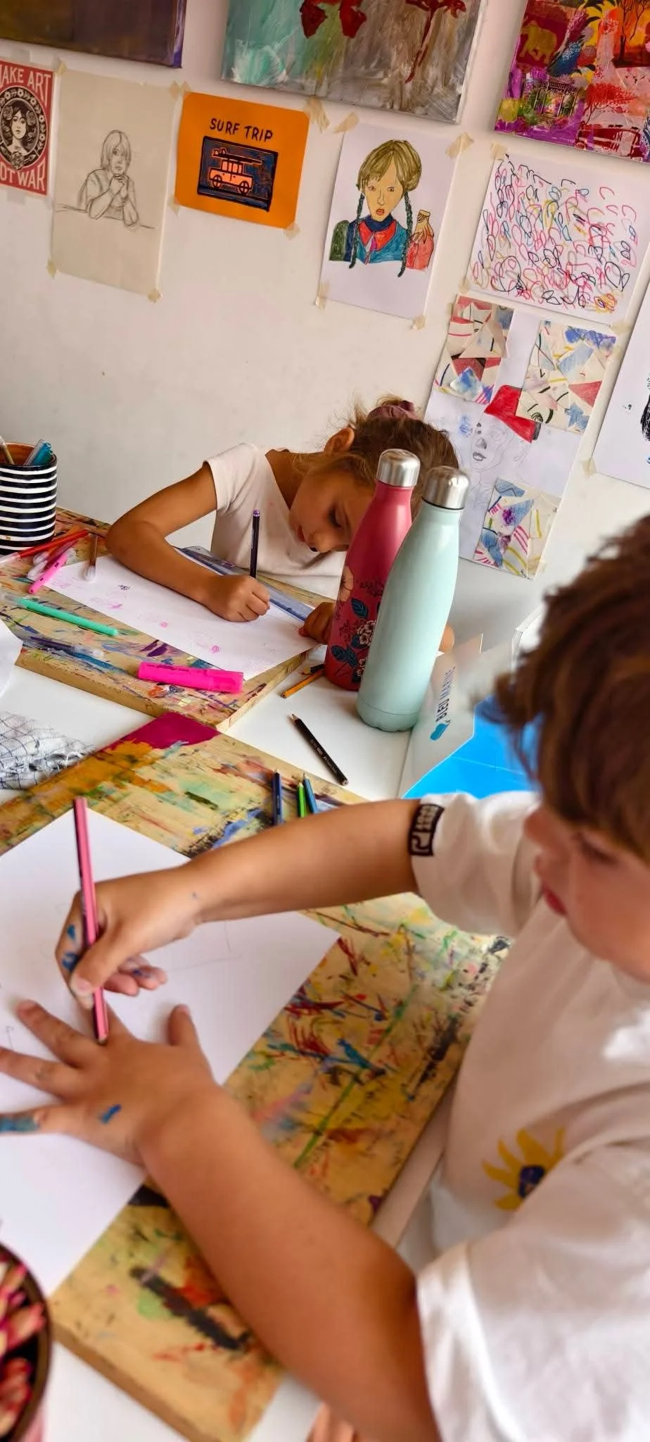 Jeune fille qui dessine avec des feutres sur une feuille blanche dans un atelier d'art, entourée de bouteilles d'eau et de matériel de dessin, avec une affiche colorée et des œuvres d'art accrochées au mur