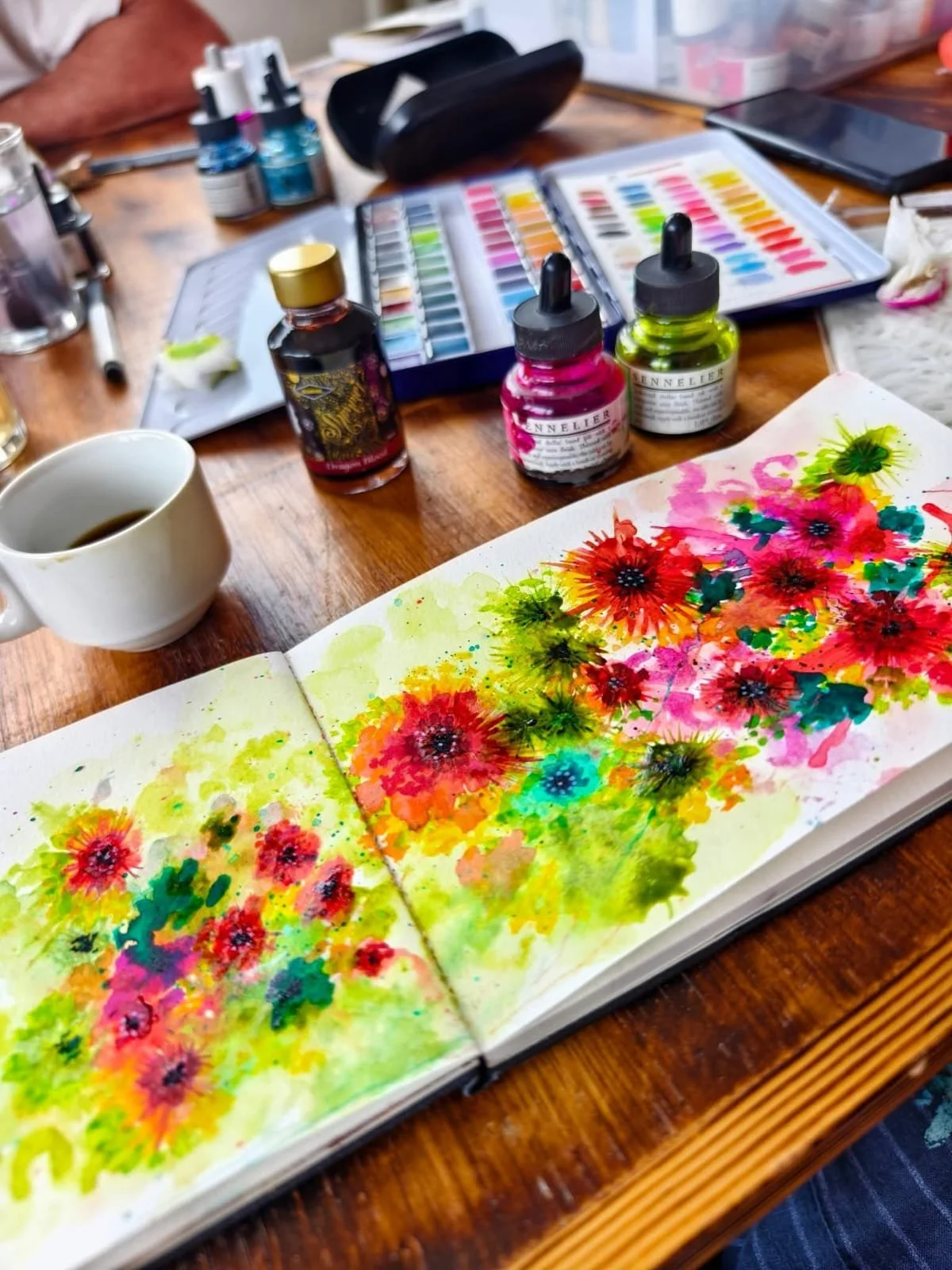 Un carnet de croquis ouvert avec une peinture de fleurs colorées, entouré de bouteilles d'encre, de pigments et d'outils de peinture sur une table en bois.