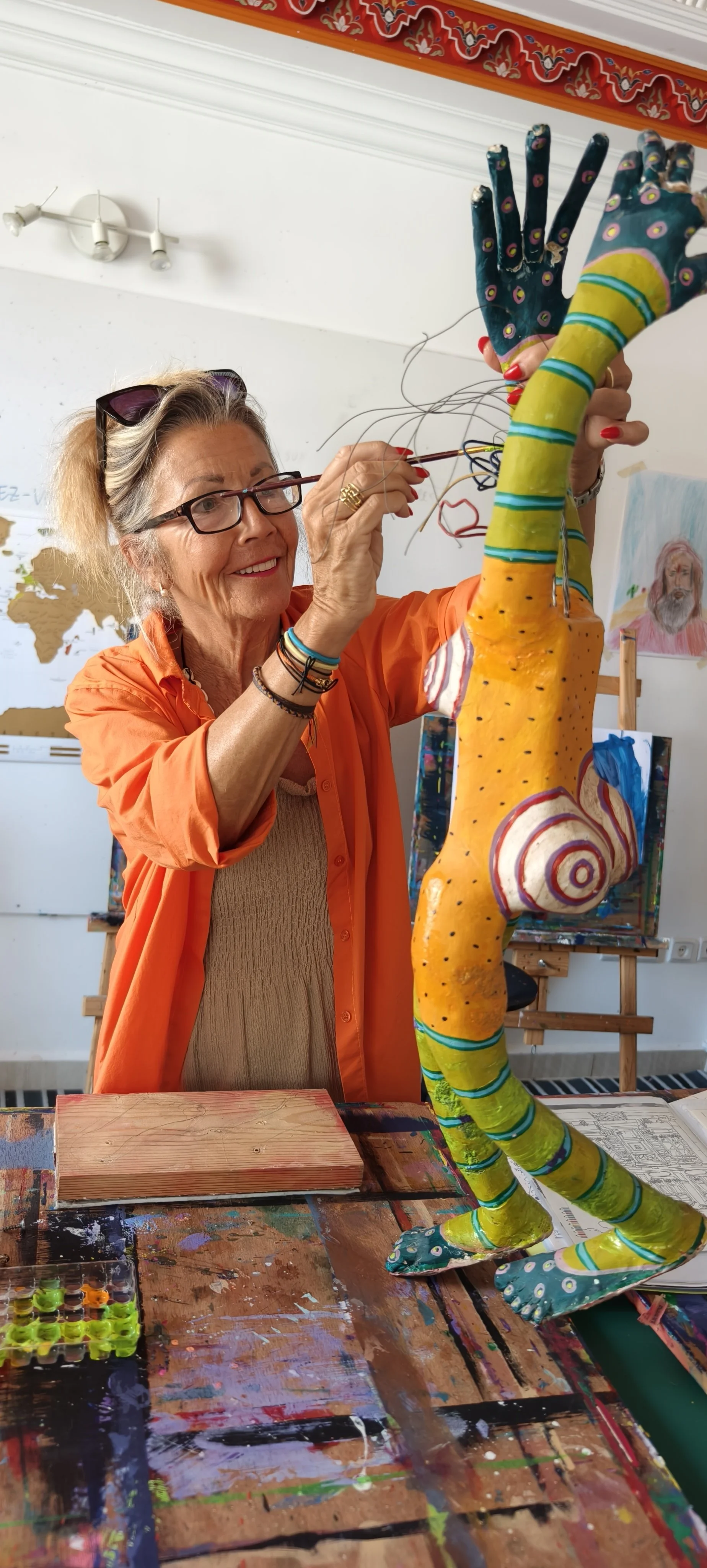Une femme âgée, portant des lunettes et une chemise orange, est en train de peindre une sculpture colorée de forme humaine dans un atelier d'art.