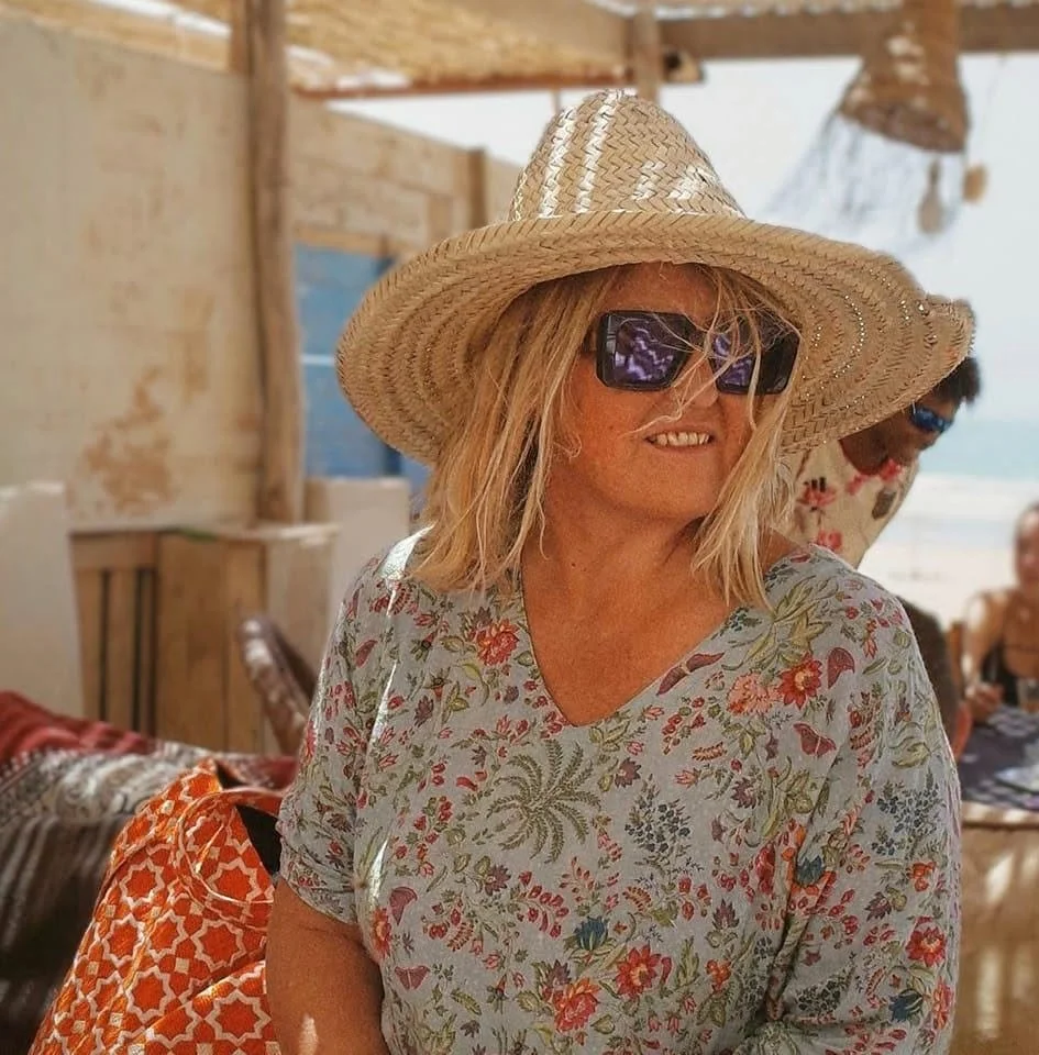 Une femme souriante porte un chapeau en paille, des lunettes de soleil et une robe à motifs floraux, dans un cadre plage en plein air.