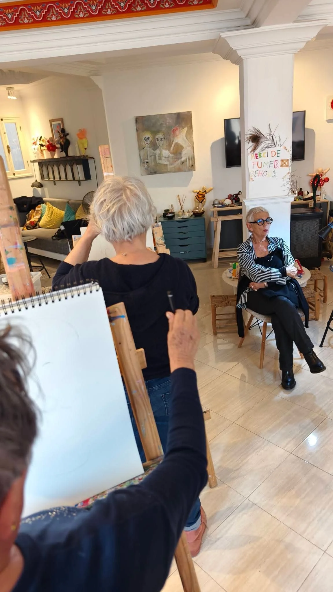 Deux femmes assises dans une pièce décorée avec des œuvres d'art et des objets de décoration, en train de discuter ou de travailler.