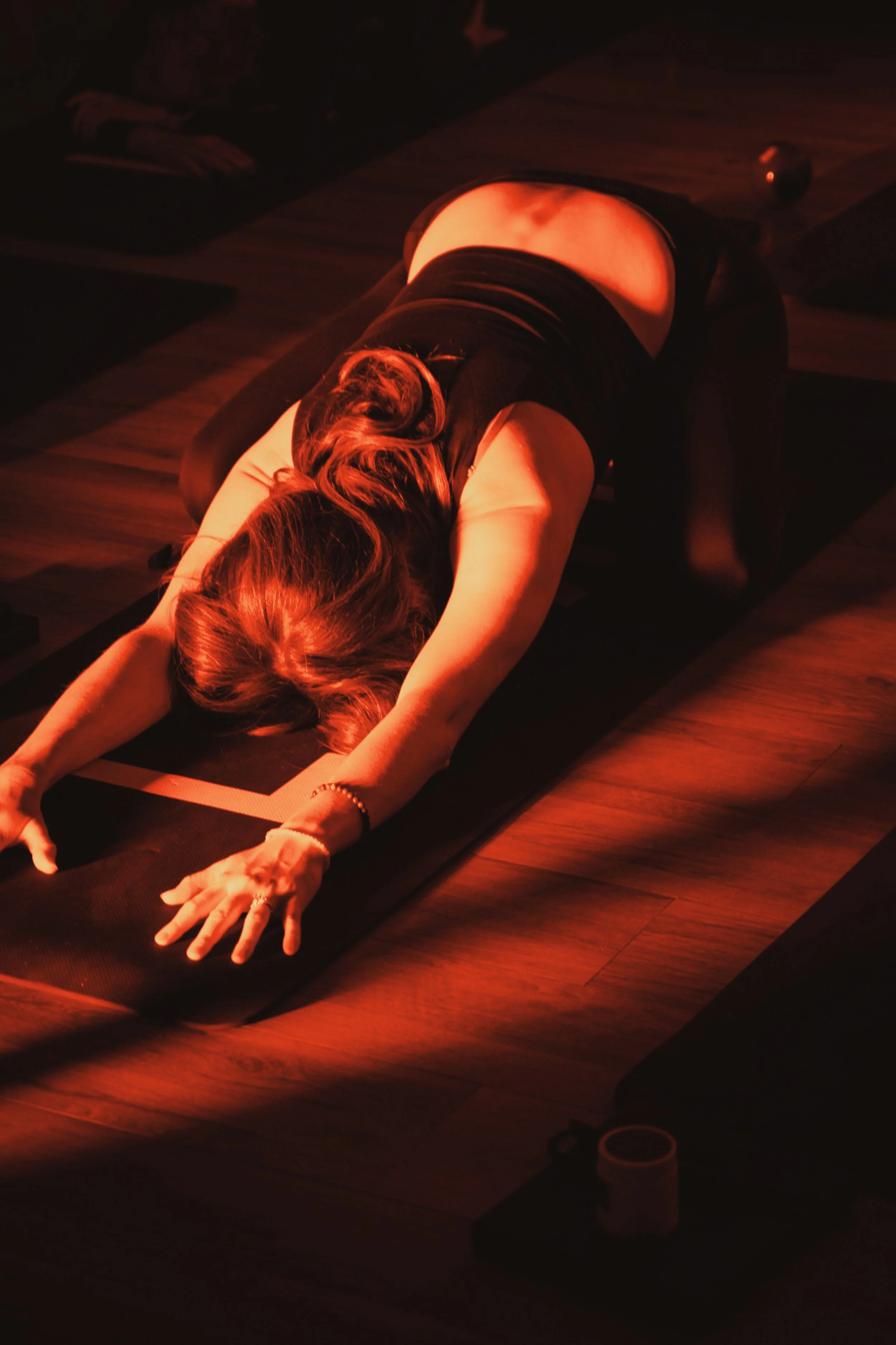 A woman practicing yoga in a dark room, wearing black workout clothes, stretching on a yoga mat under warm red lighting.