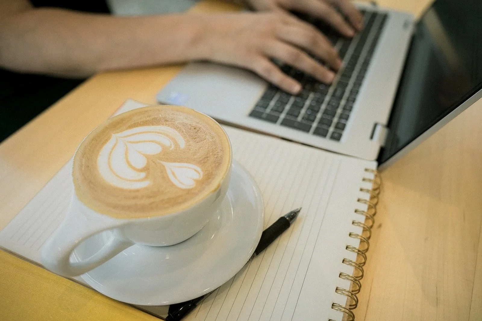 A cup of latte with latte art on top, a black pen, an open lined notebook, a silver laptop, and a person's hand on the laptop keyboard on a wooden table.