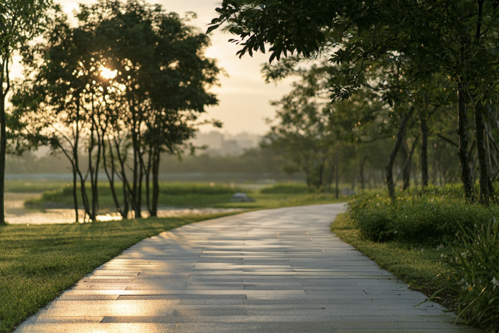 A paved walking trail winding through a park with green grass, trees, and a water feature in the background during sunset.