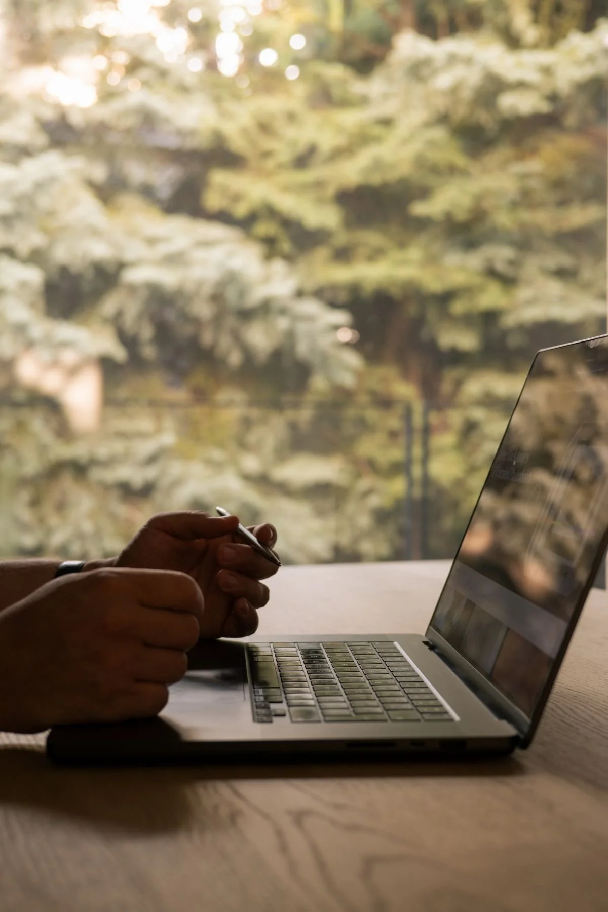 Person holding a pen over a laptop with a blurred green outdoor background.