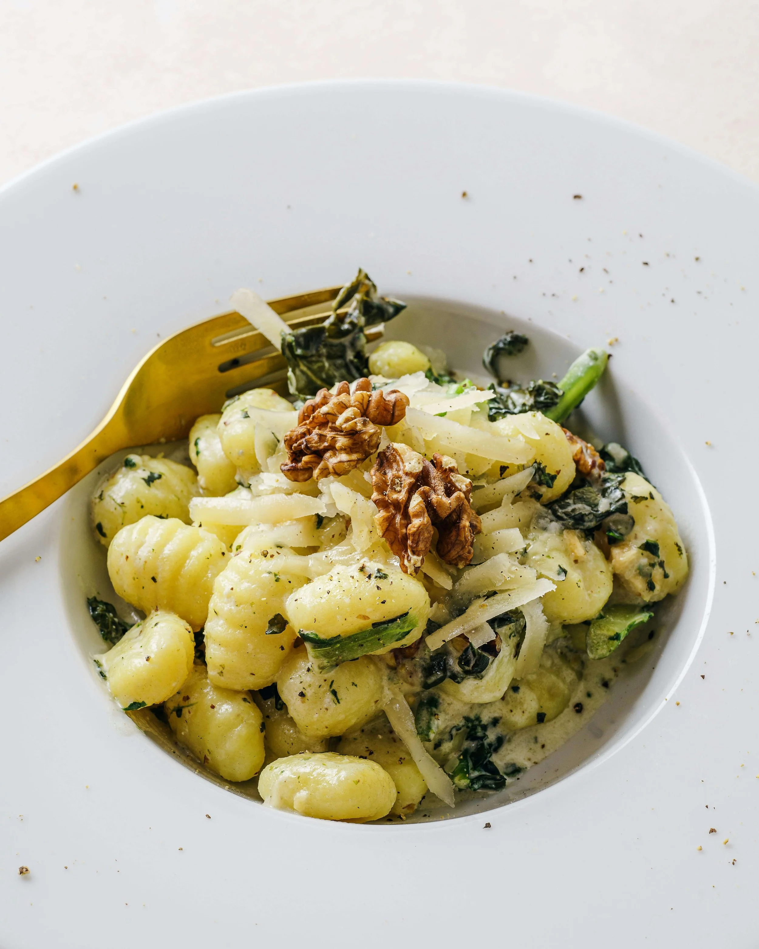 Close-up of a white bowl filled with gnocchi pasta covered in a creamy sauce, topped with walnuts and grated cheese, with black pepper and herbs sprinkled around.