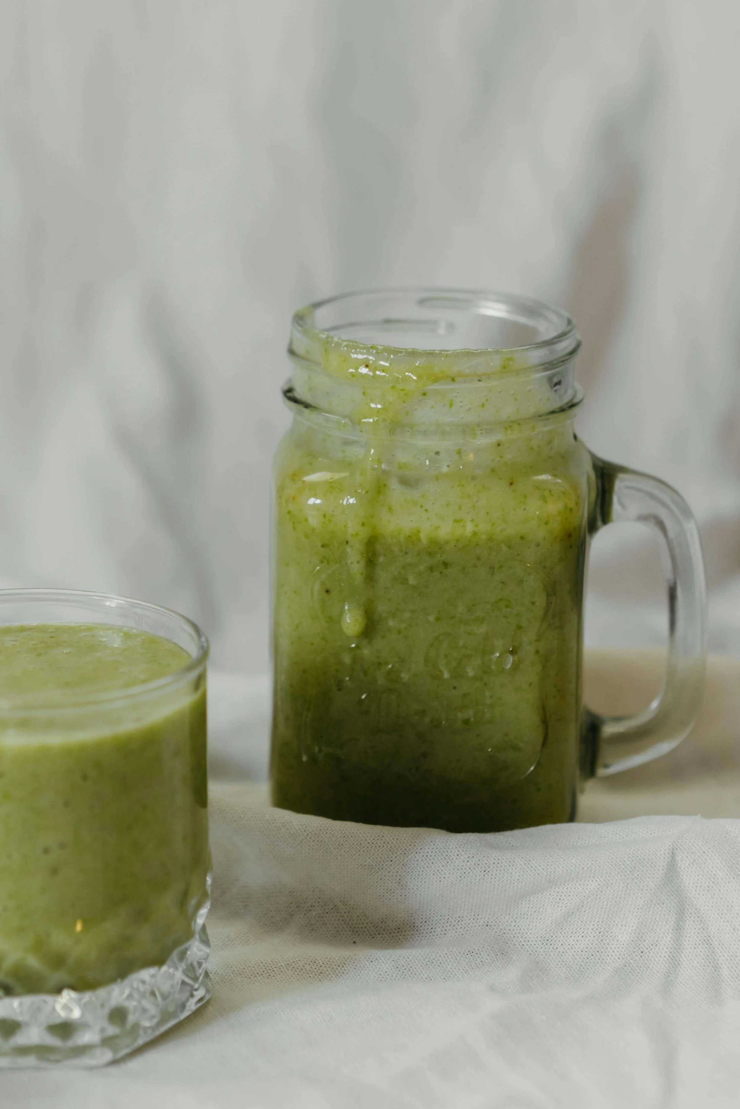 Close-up of a mason jar and a glass cup filled with green smoothie on a white surface against a white background.