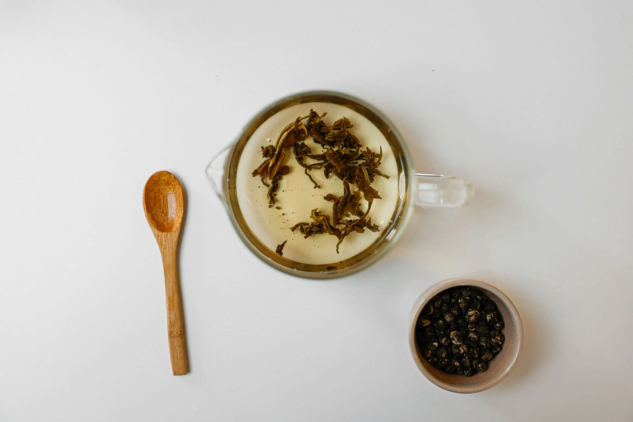 Top-down view of a glass cup with dried herbs inside, a small bowl of black peppercorns, a small white bowl with black salt, and a wooden spoon on a white surface.