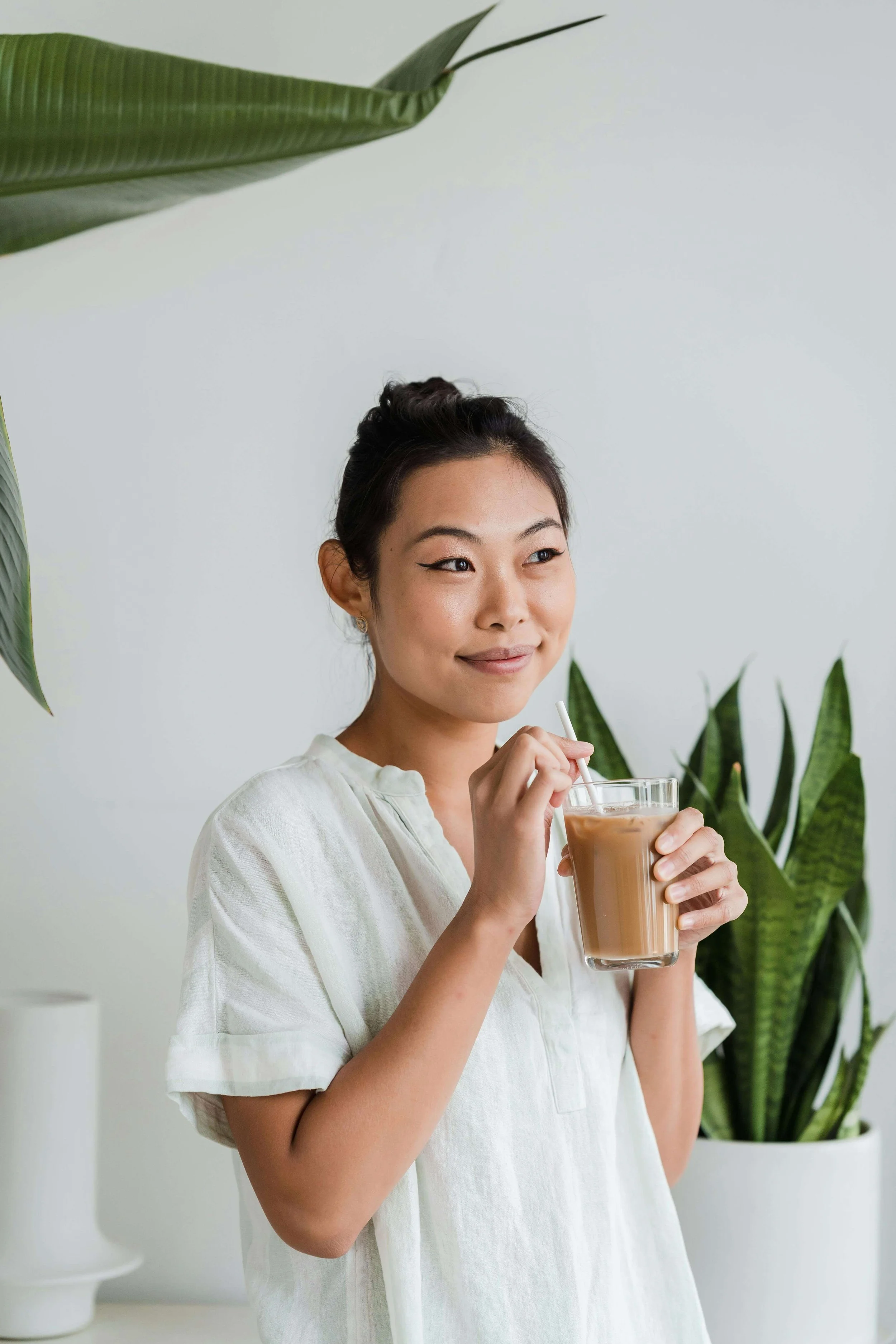A young woman with dark hair in an updo, wearing a light-colored top, is smiling and holding a glass of iced coffee or tea with a straw, standing in front of a white wall with large green houseplants.