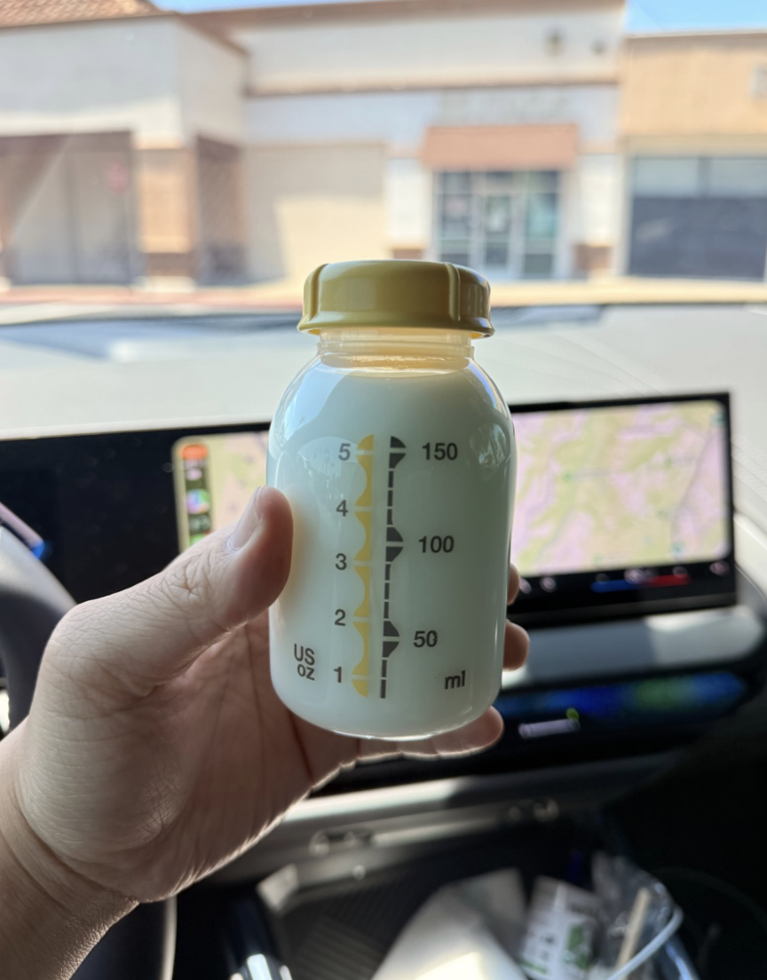 Hand holding a baby bottle with measurement markings in front of a car dashboard and a GPS navigation screen.
