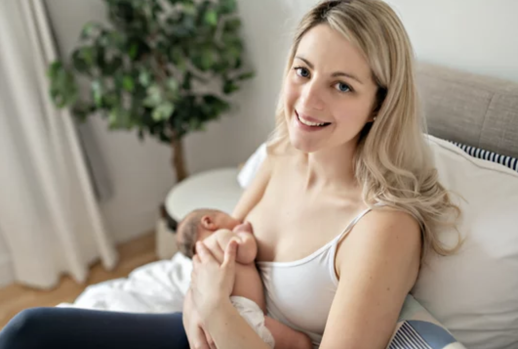 A smiling woman breastfeeding her baby while sitting on a bed in a cozy, well-lit room with houseplants and curtains.