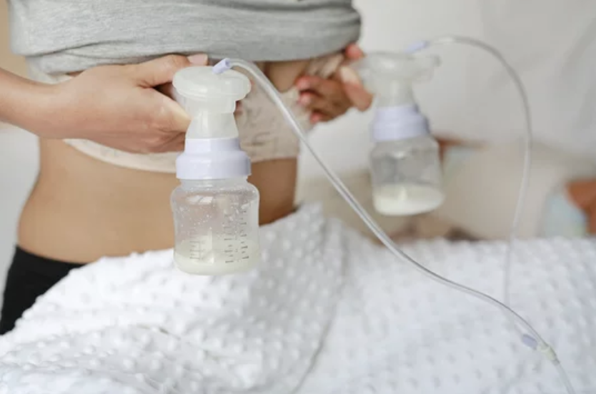 Person holding a breast pump with milk collection bottles attached, positioned near a baby lying on a white textured blanket.